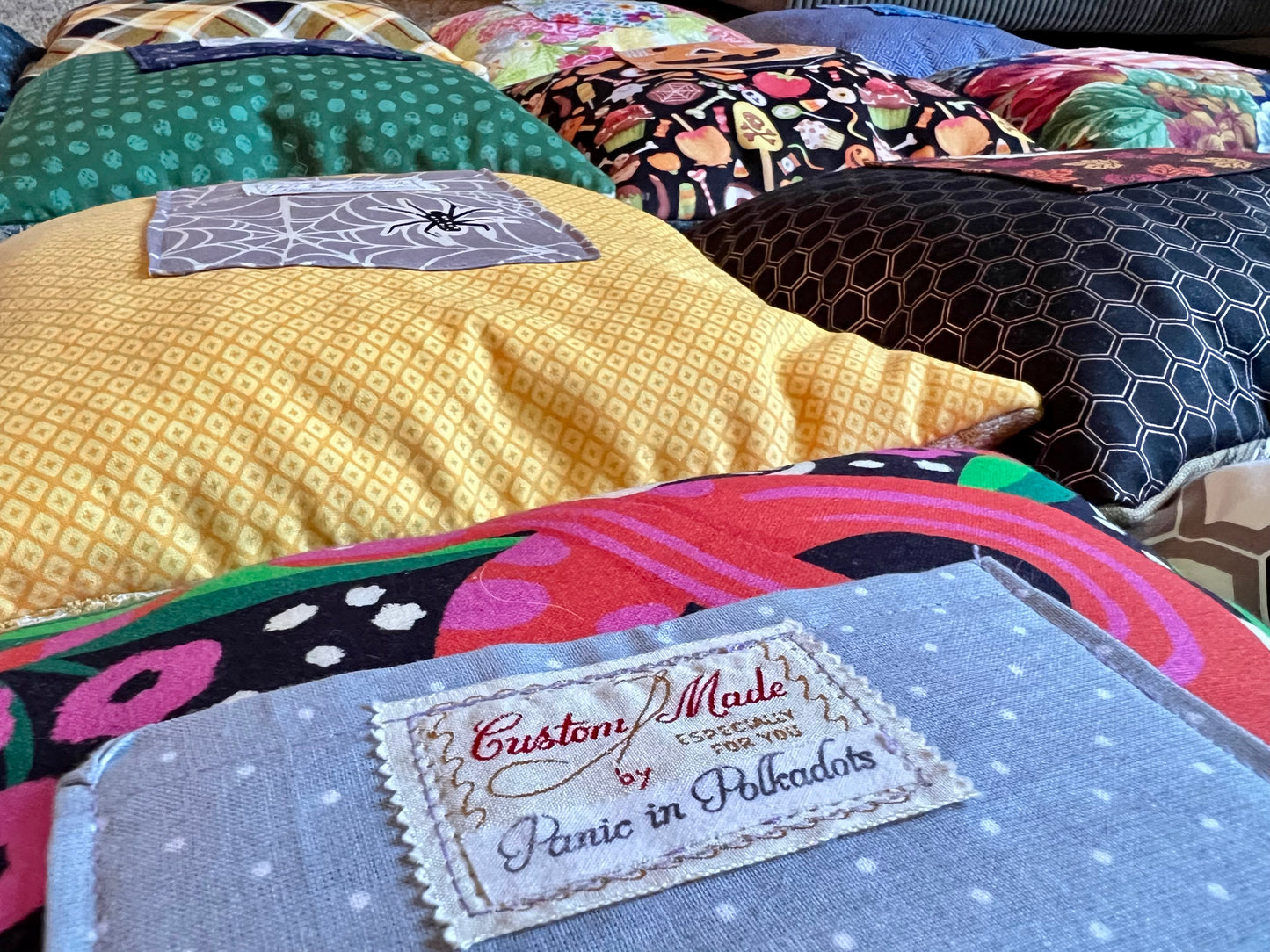 A close-up of a handmade quilted cushion in a light blue polka-dot fabric with a custom patch reading 'Custom Made by Panic in Polkadots,' set against a background of other colorful patterned cushions.