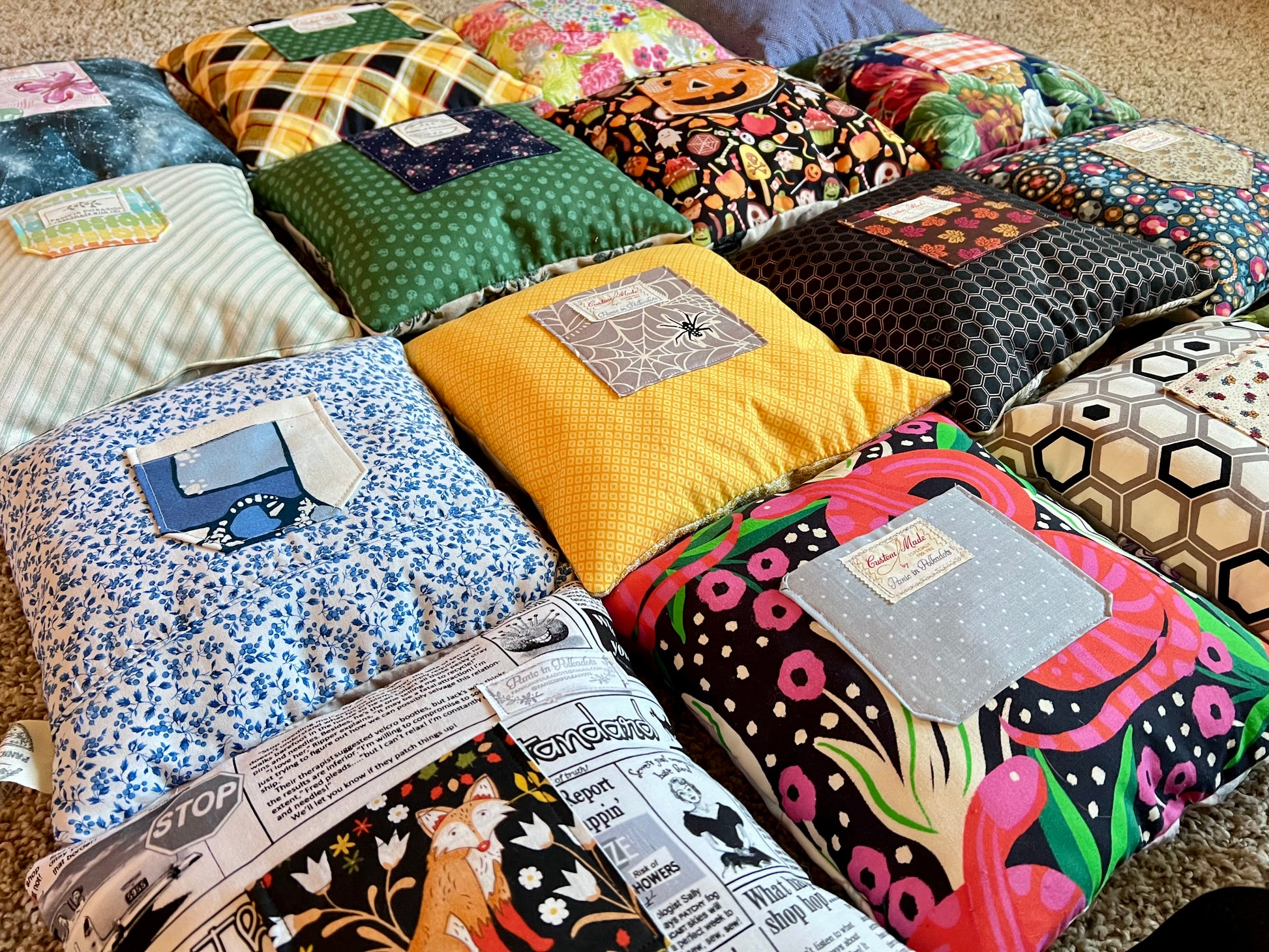 A collection of handmade, patchwork-style decorative pillows arranged on a carpeted floor. The pillows feature a variety of colorful fabrics, including floral prints, geometric patterns, newspaper clippings, and solid colors.