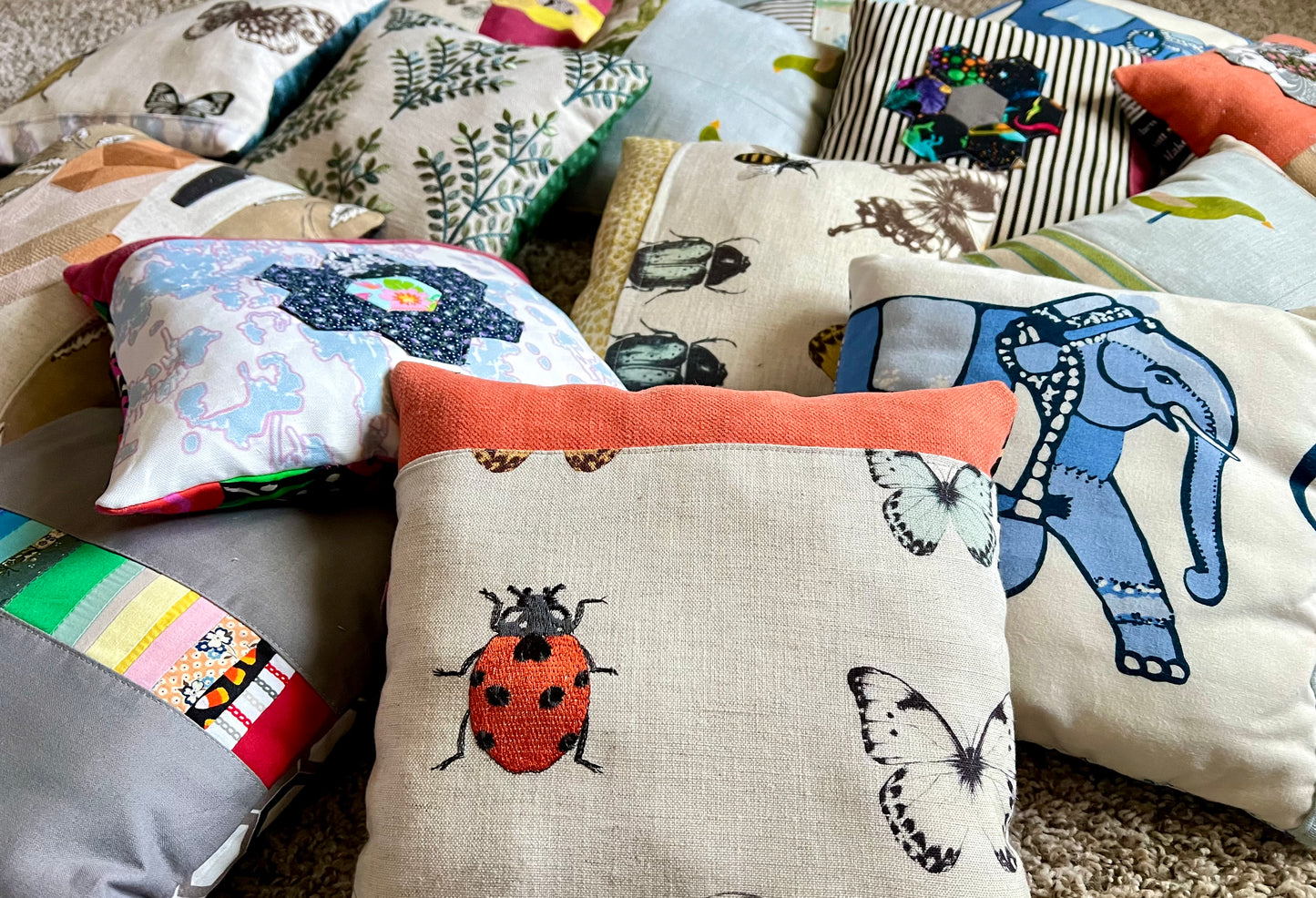 A collection of handmade, multi-colored fabric throw pillows with various animal and nature prints, including a prominent central beige pillow with a red ladybug and butterfly pattern.
