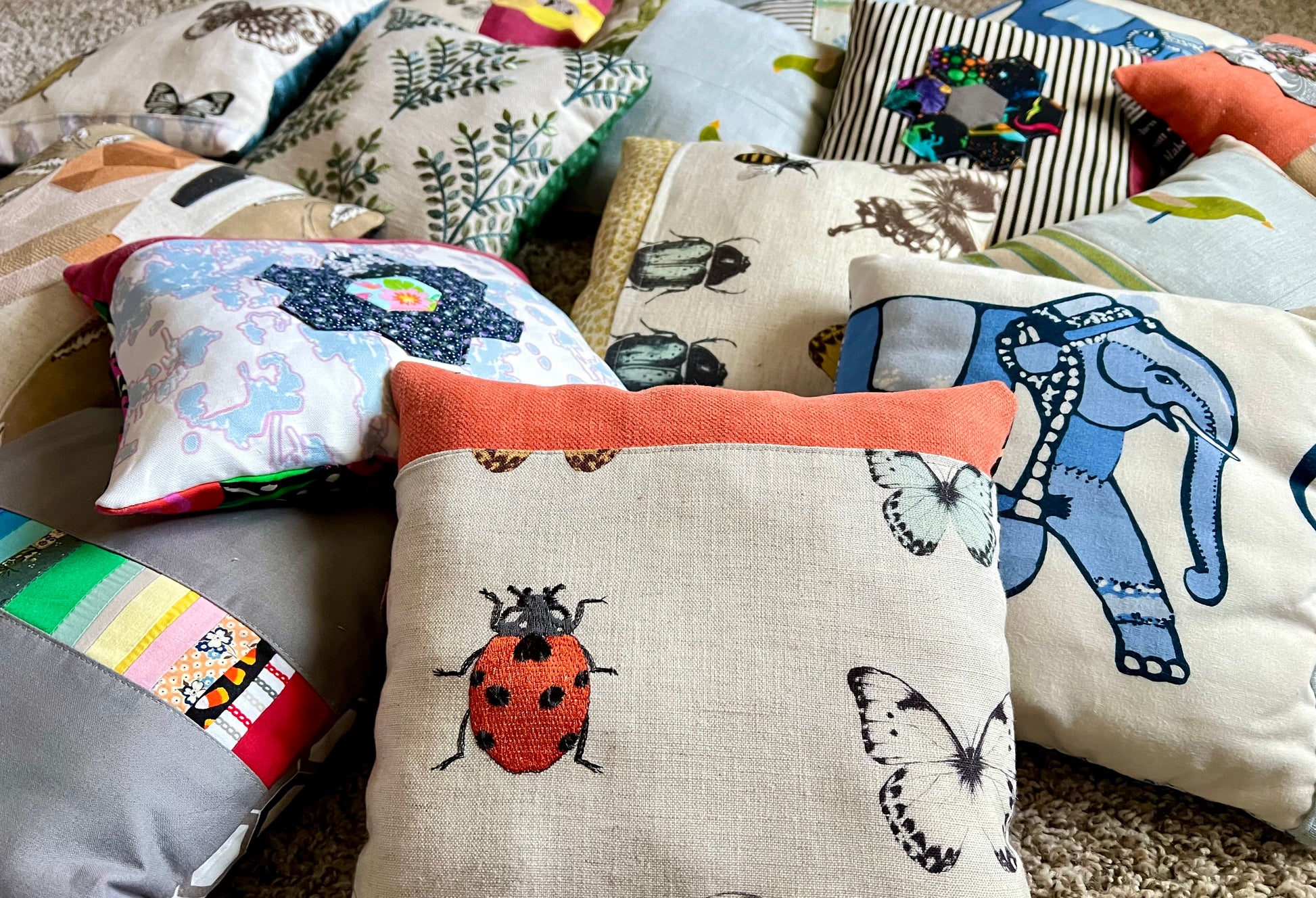 A collection of handmade, multi-colored fabric throw pillows with various animal and nature prints, including a prominent central beige pillow with a red ladybug and butterfly pattern.