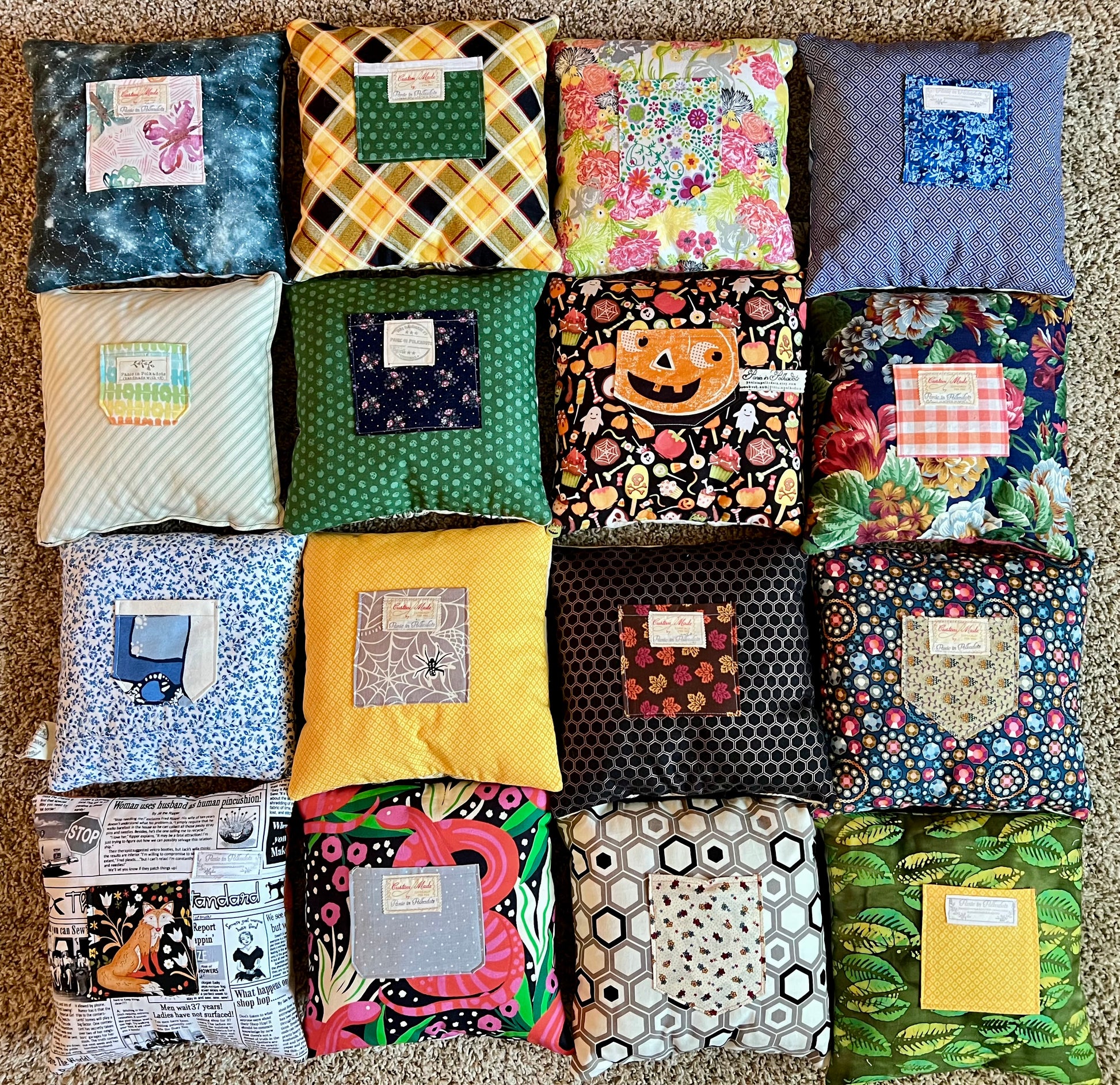 A grid of 15 square fabric pillows arranged on a beige carpet, each with a different colorful printed fabric exterior and a small white label patch on the front of each tooth fairy pocket.