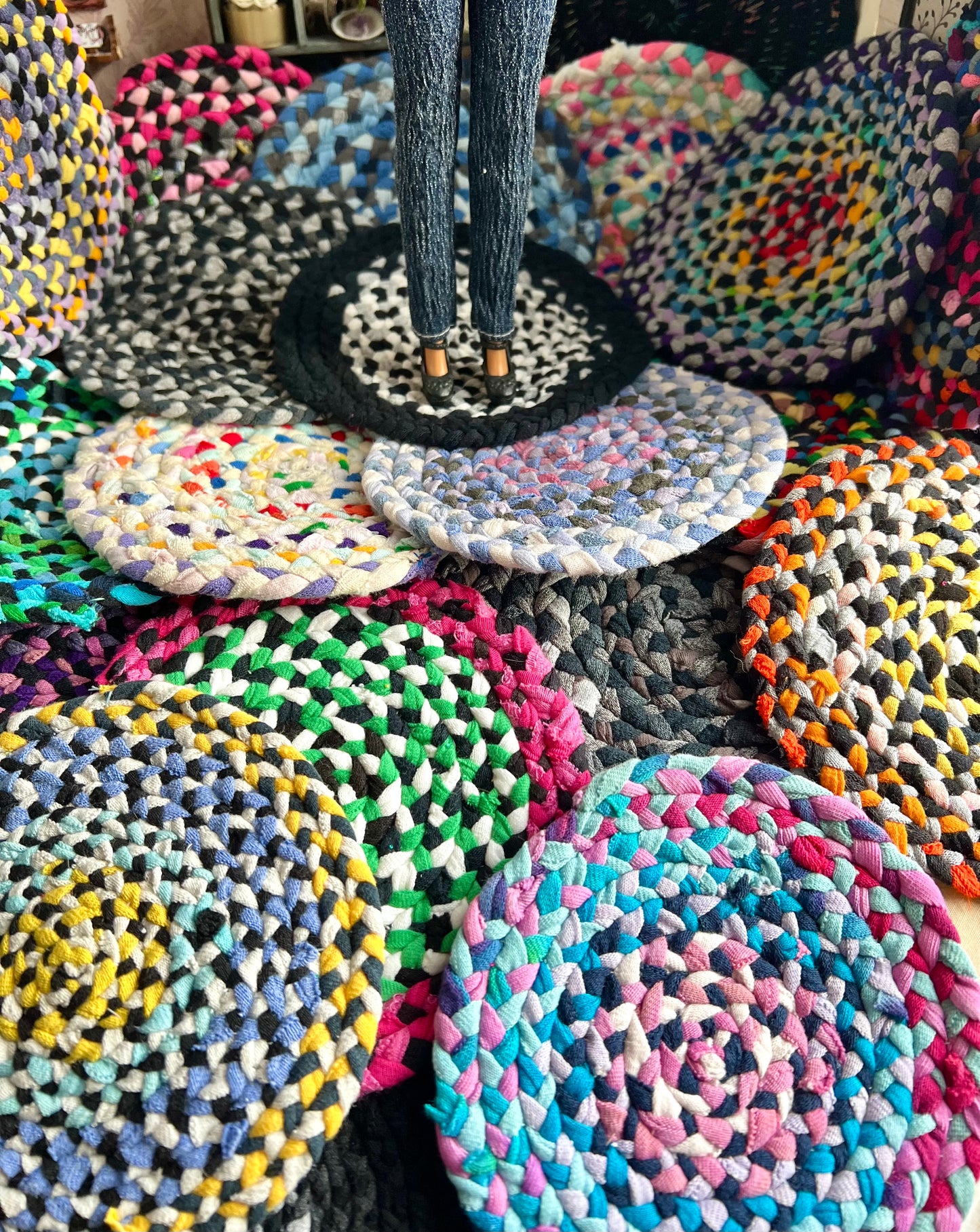 Colorful braided miniature rugs arranged on a surface with a doll standing on one of them.