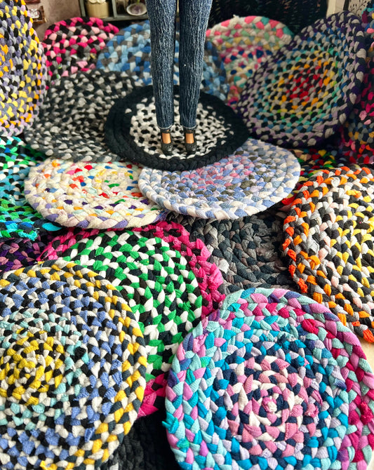 Colorful braided miniature rugs arranged on a surface with a doll standing on one of them.
