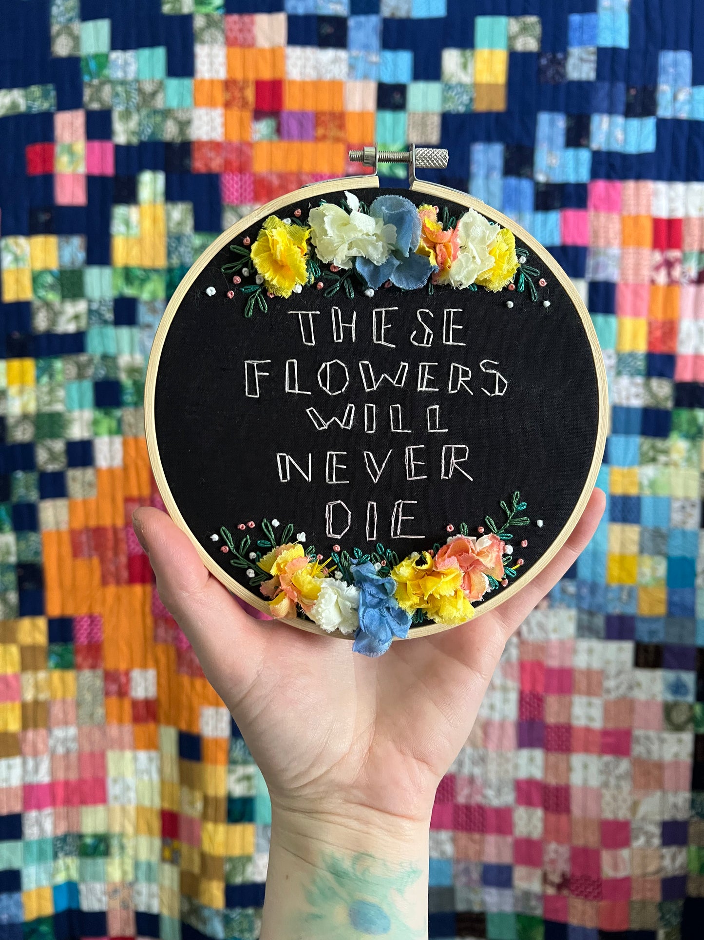 Embroidery these flowers will never die, held in hand for scale, with colorful quilt in background