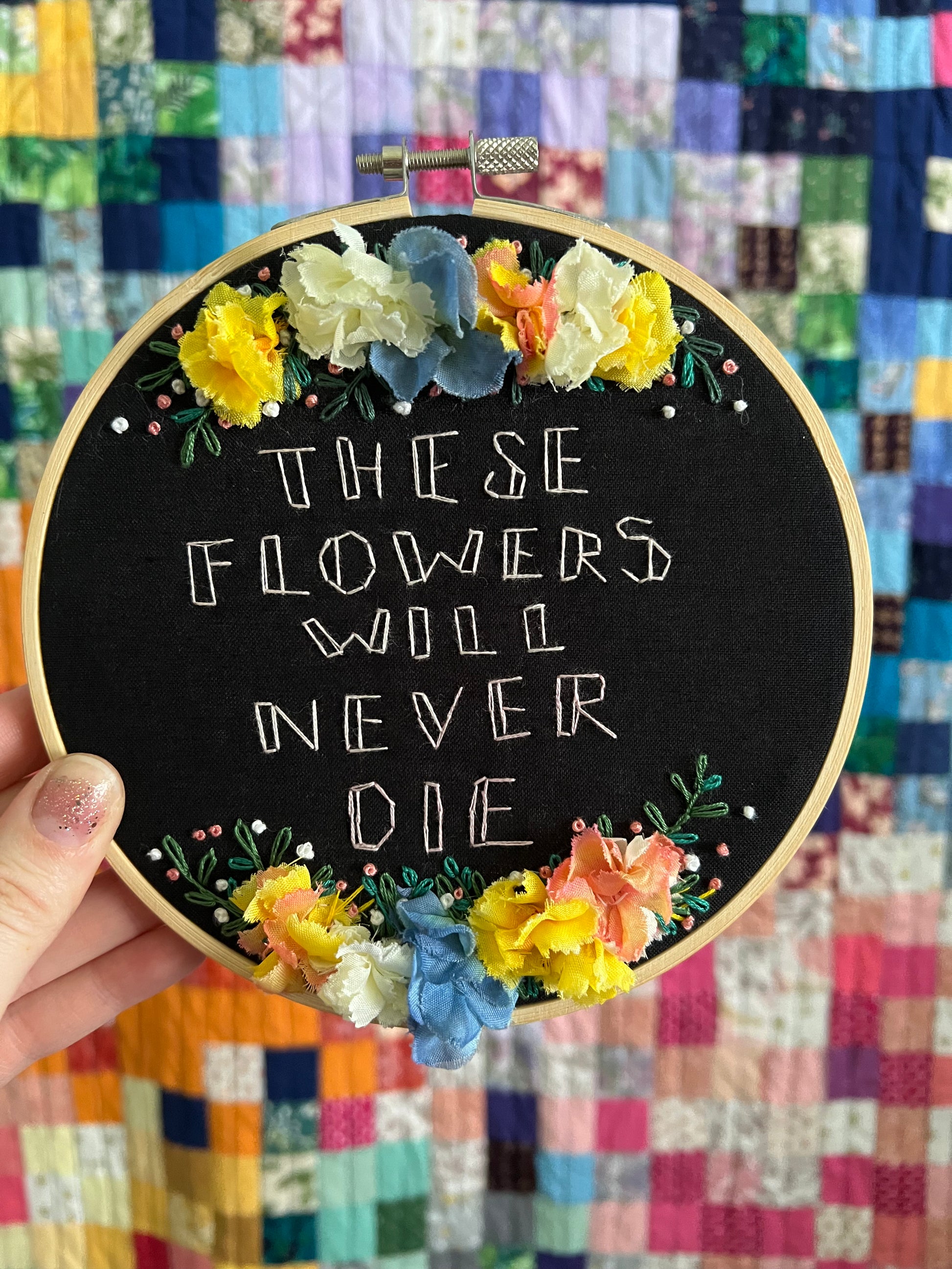 Embroidery these flowers will never die, held in hand for scale, with colorful quilt in background