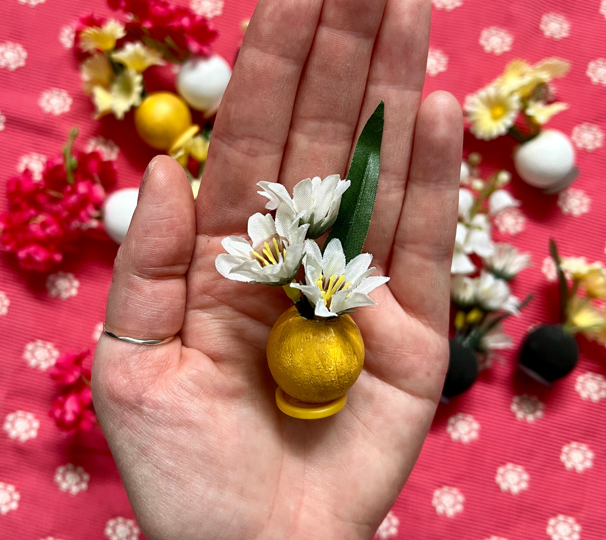 Small decorative item resembling a yellow vase with white flowers held in a hand against a red background with floral patterns.