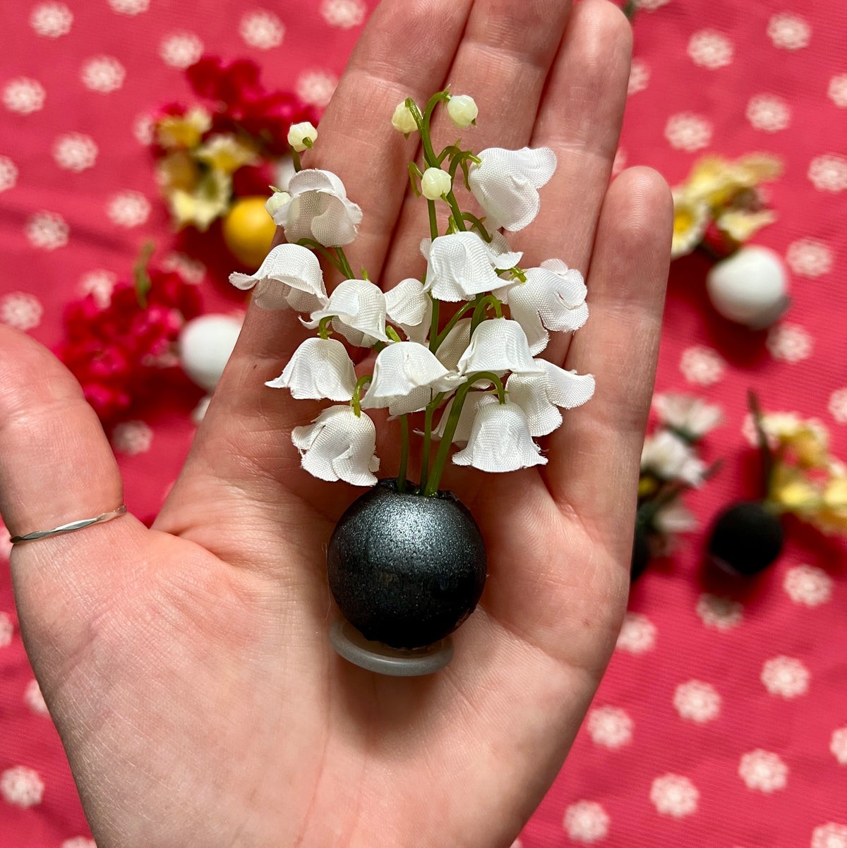 Hand holding a small black vase with white flowers against a red polka dot background