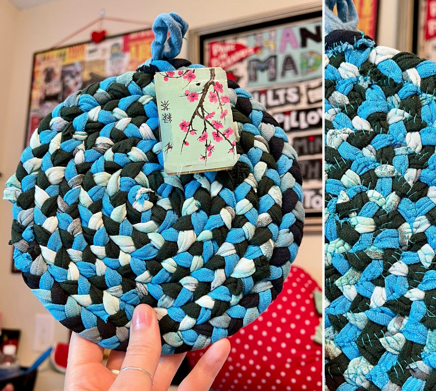 A round braided fabric trivet or pot holder made from mixed blue, black, and white denim scraps, held by a hand in an indoor setting. A light green paper tag with a cherry blossom illustration is attached to the top.