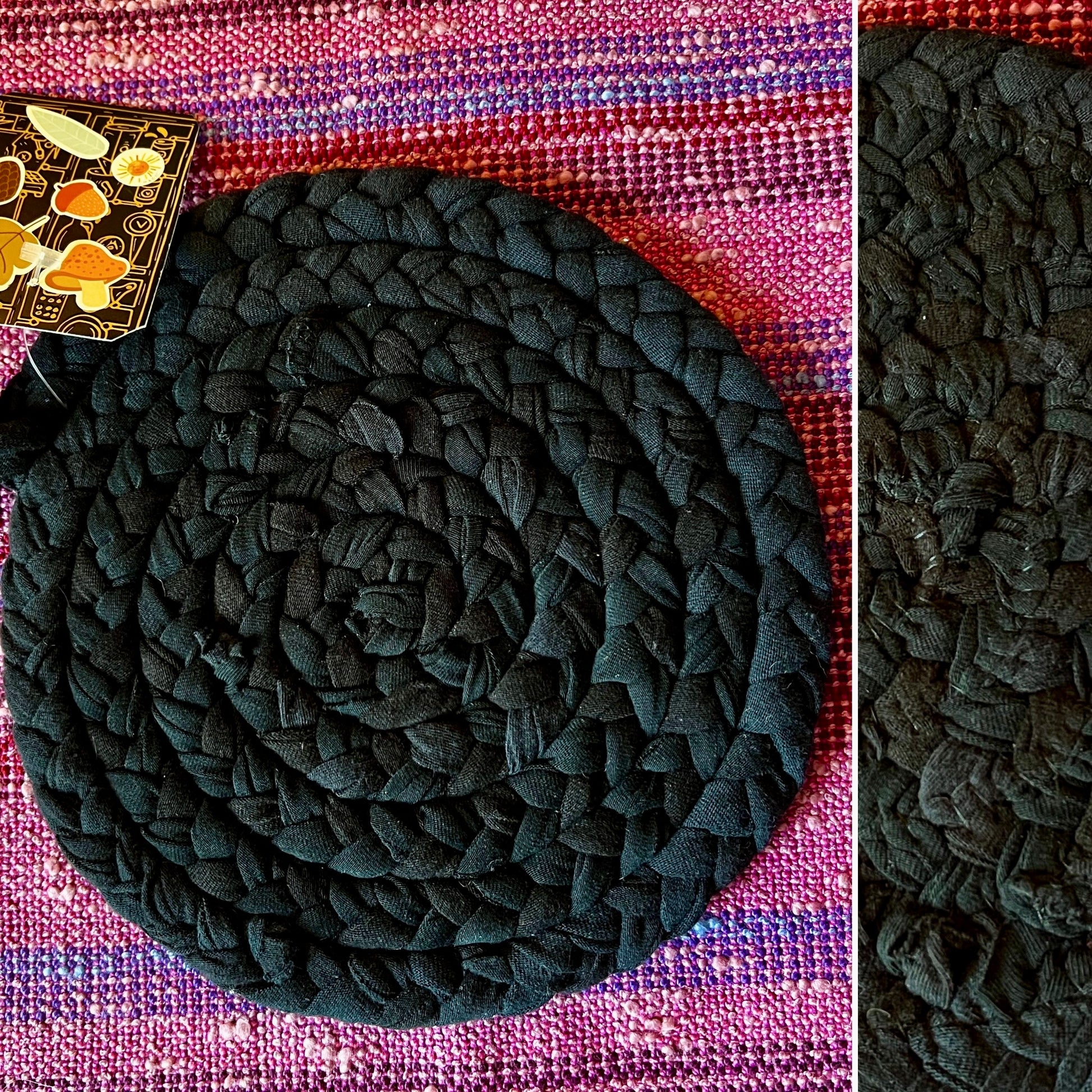 A round braided rug or mat in very dark charcoal/black, shown on a woven pink and purple textile background with a small decorative tag visible in the upper left. A narrow side panel on the right shows a close-up of the braided and hand-sewn texture.