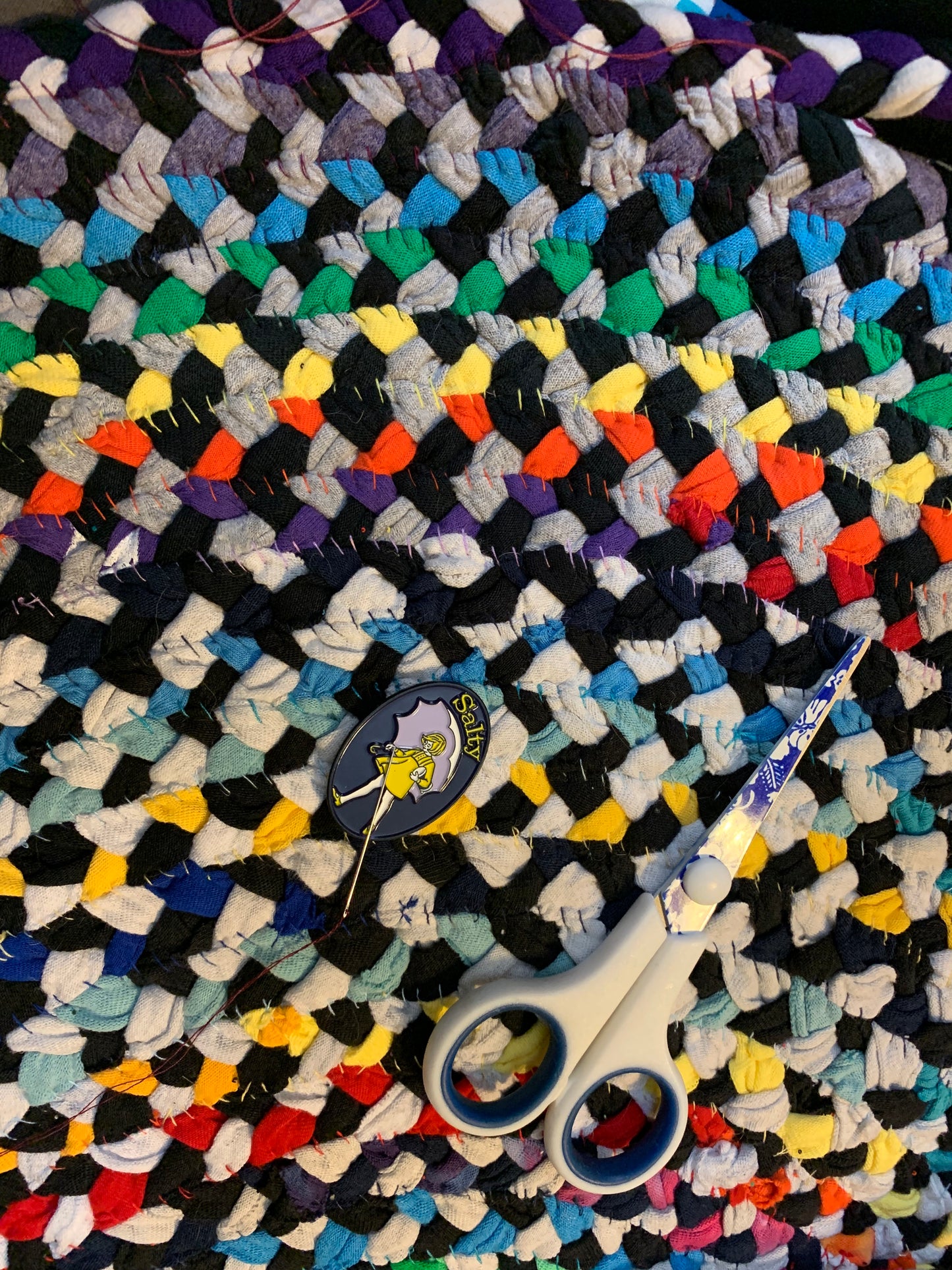 Rug in progress, pictured with a pair of scissors, a needle minder holding a needle with purple thread. The back stitches are coordinating with the braid color. 