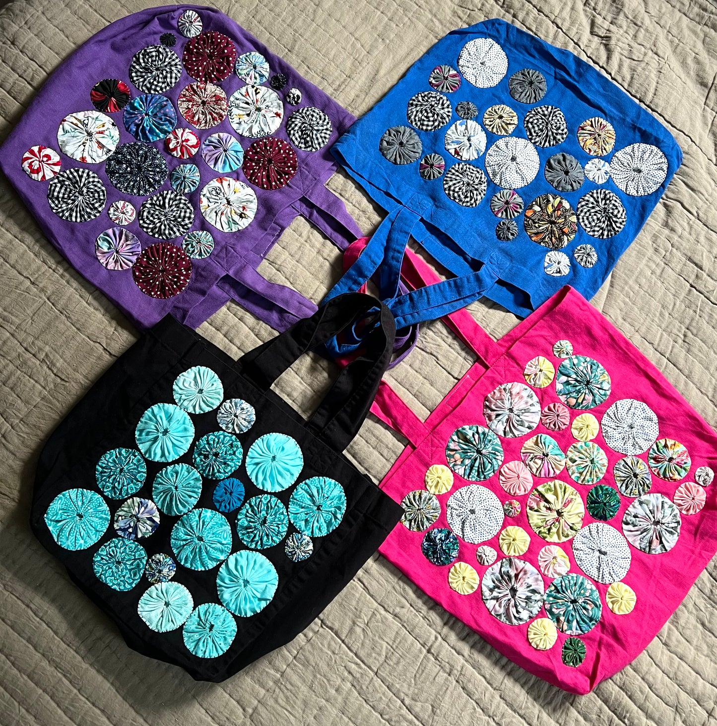 a collection of four tote bags, in various colors, with handsewn yoyos on the front of each one. The handles are layered together in the center