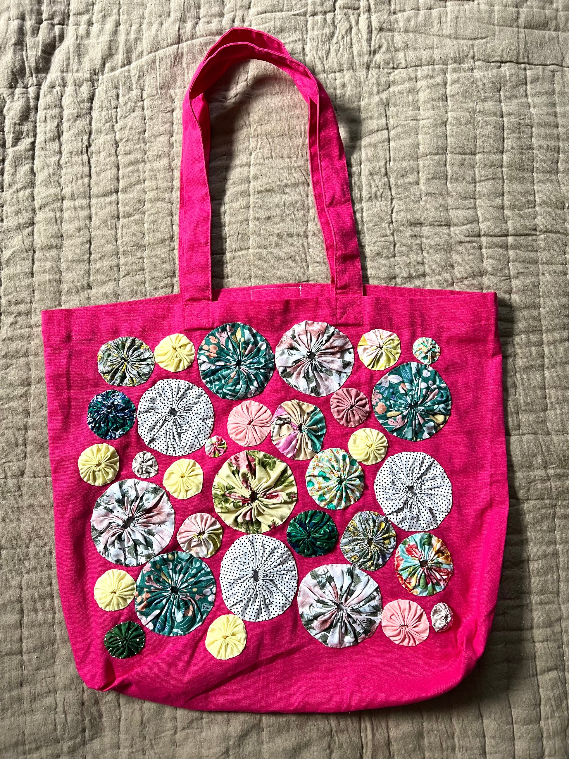 pink tote bag with white, green and yellow yoyo circles sewn on top, aerial view against a textured quilt background