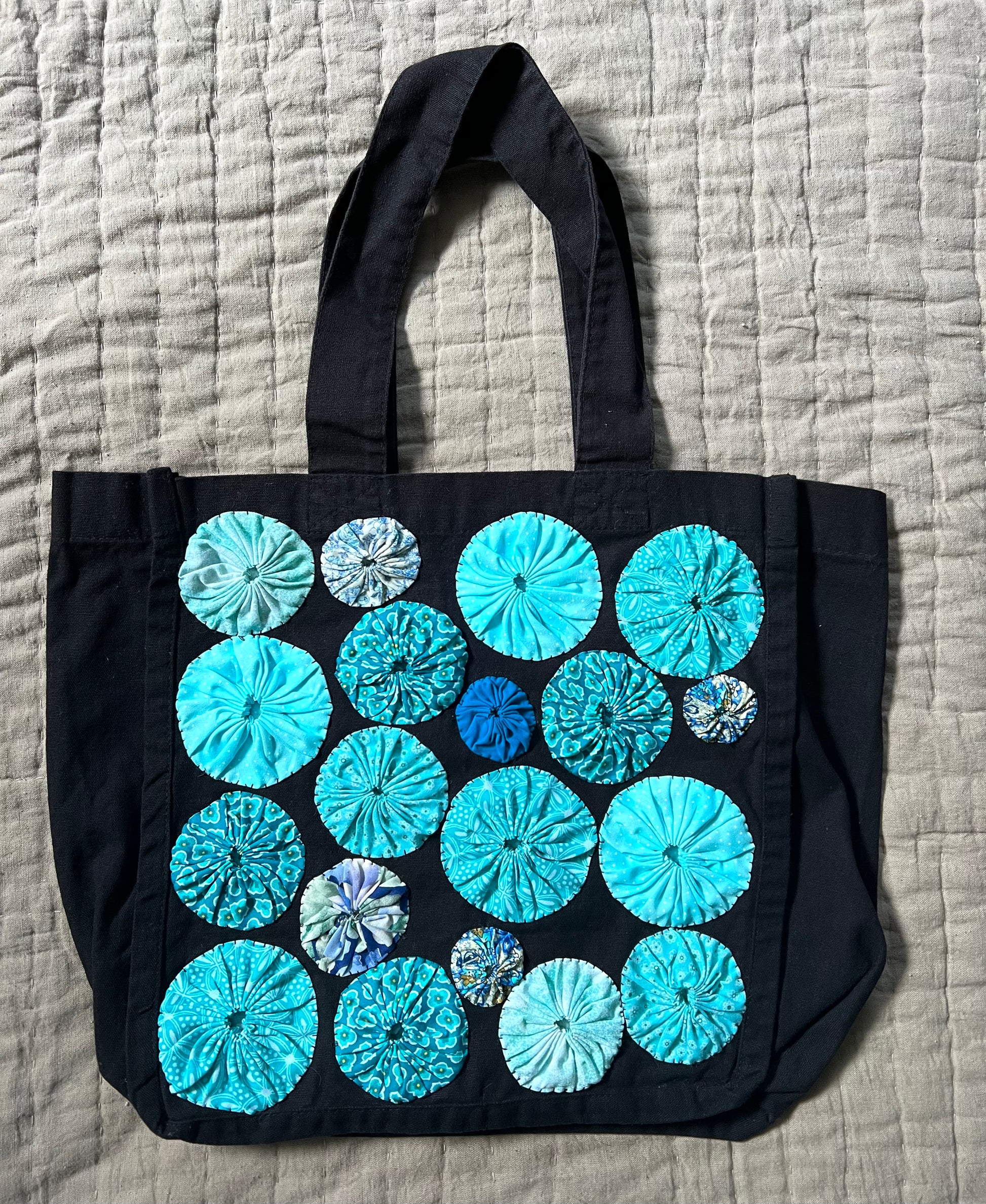 black tote bag with light blue yoyo circles sewn on top, aerial view against a textured quilt background