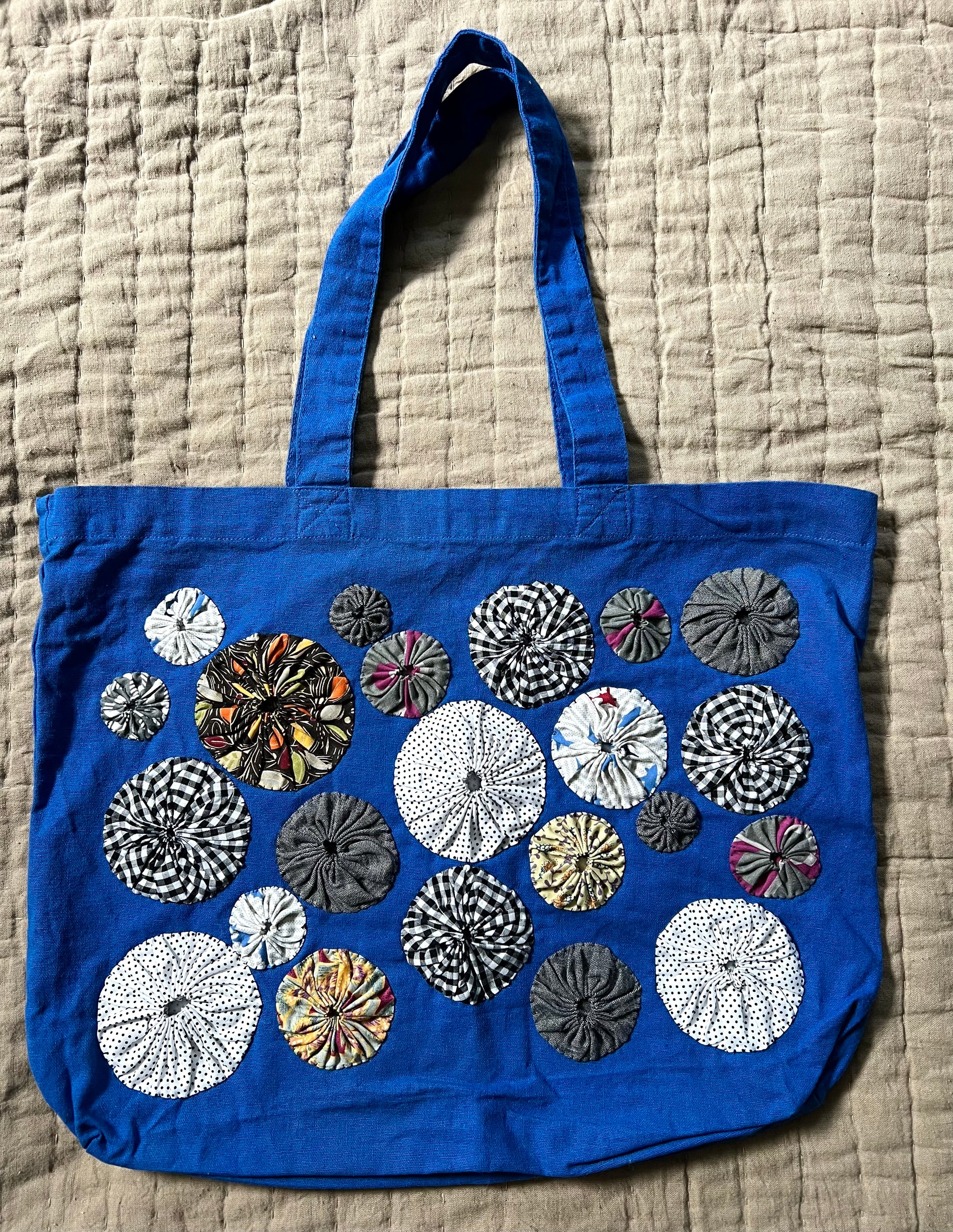 royal blue tote bag with black and white patterned and grey yoyo circles sewn on top, aerial view against a textured quilt background