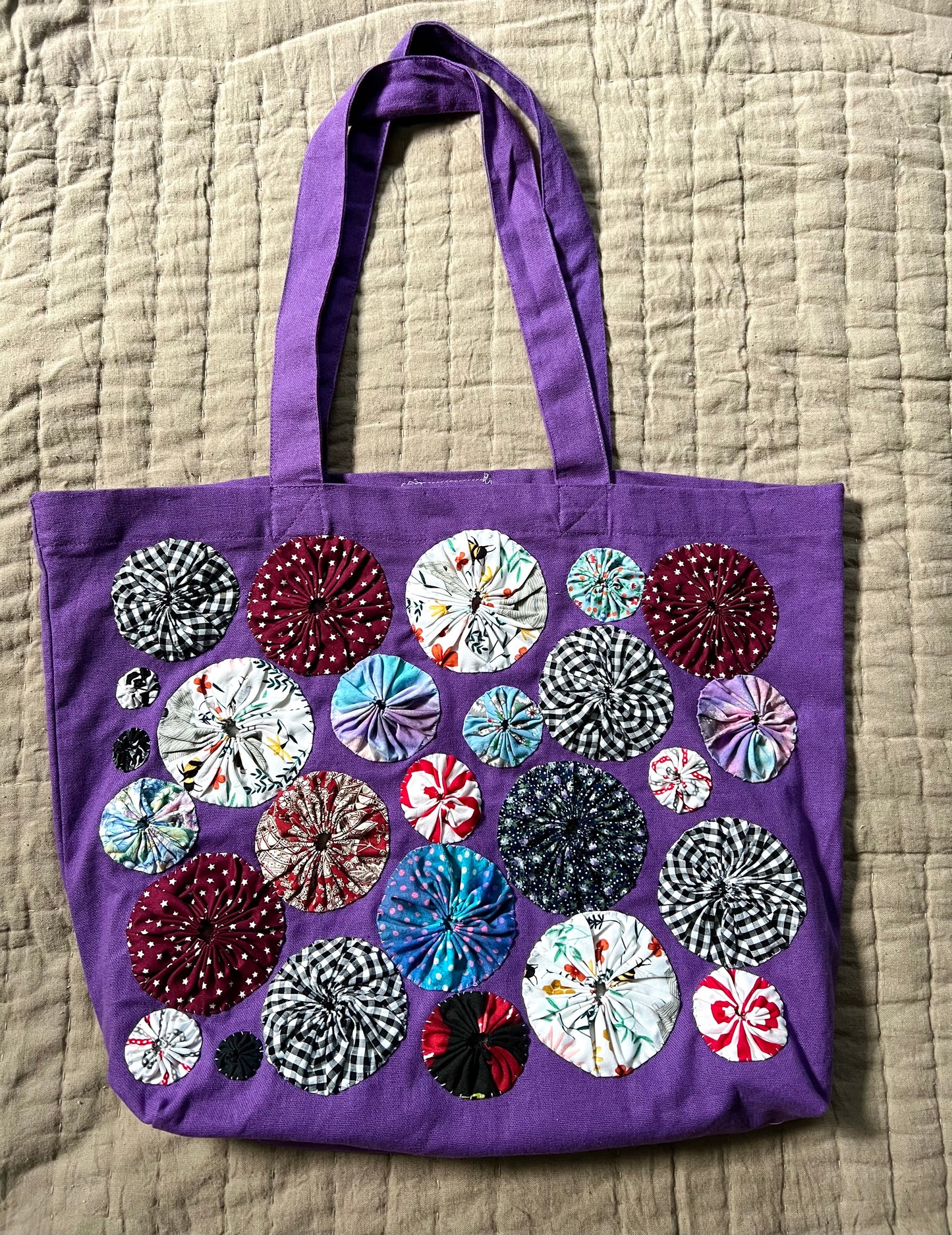 purple tote bag with red, maroon and black and white patternedyoyo circles sewn on top, aerial view against a textured quilt background