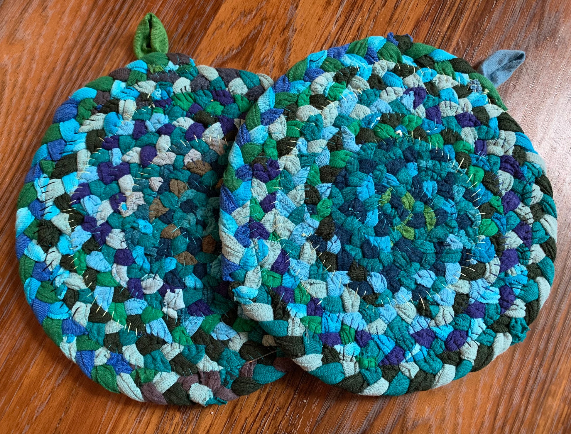 The back of two trivet potholders, to show off stitching, lay flat on a wood surface.