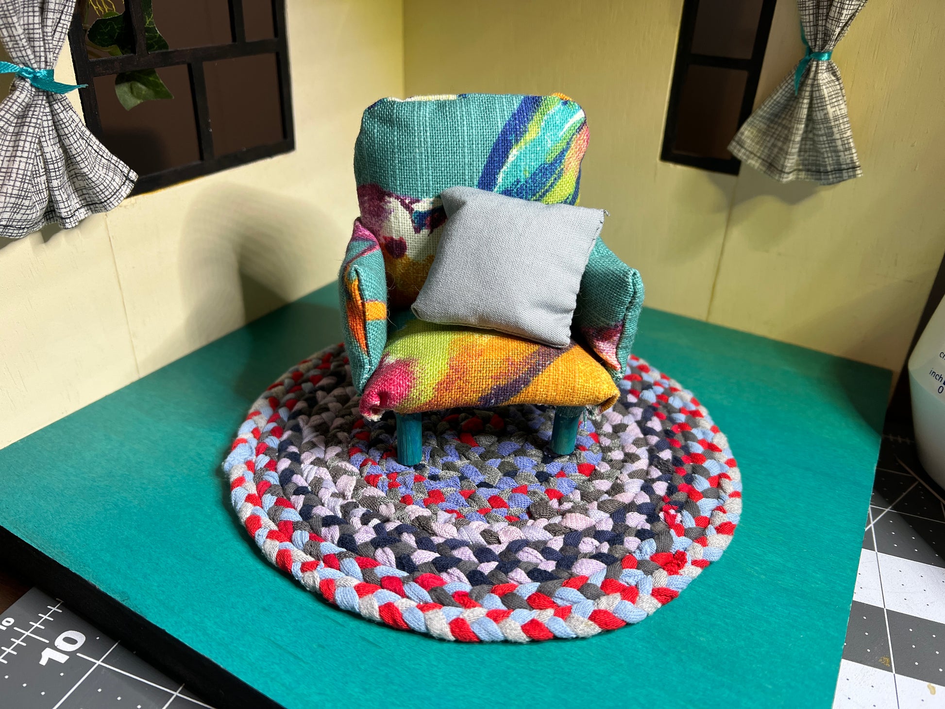 a miniature handmade dollhouse chair, on top of a mini rug (sold seperately) in a dollhouse room box. This one is a colorful teal and yellow chair with birds on it and a light grey accnt pillow