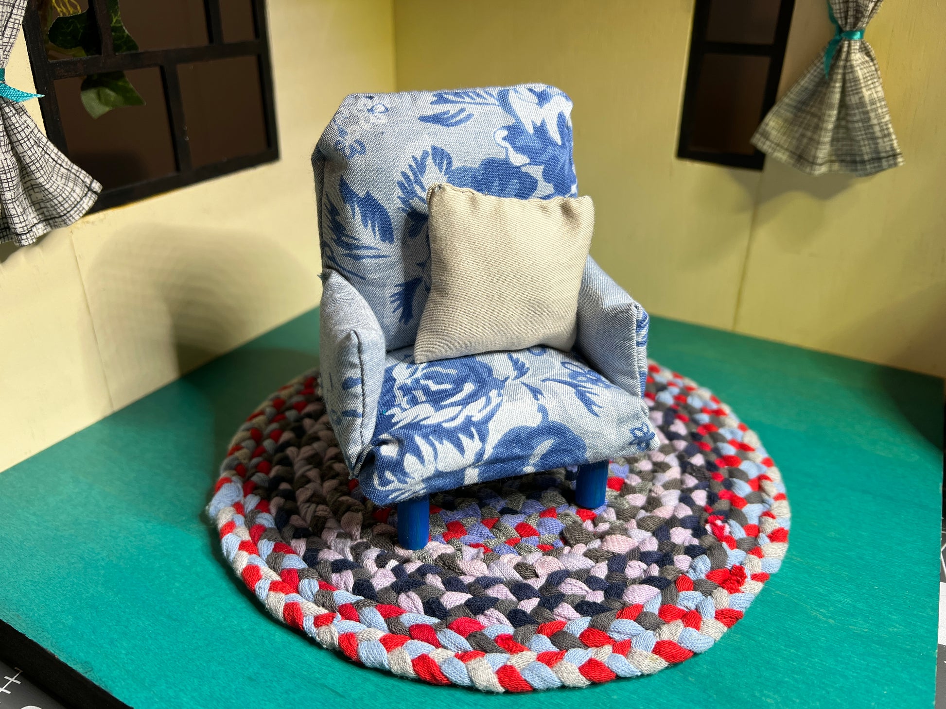 a miniature handmade dollhouse chair, on top of a mini rug (sold seperately) in a dollhouse room box. This one is a blue floral chair with a grey accent pillow