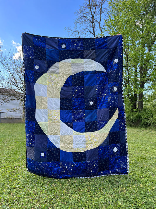 a crescent moon quilt is held up outside, against a green background with blue skies