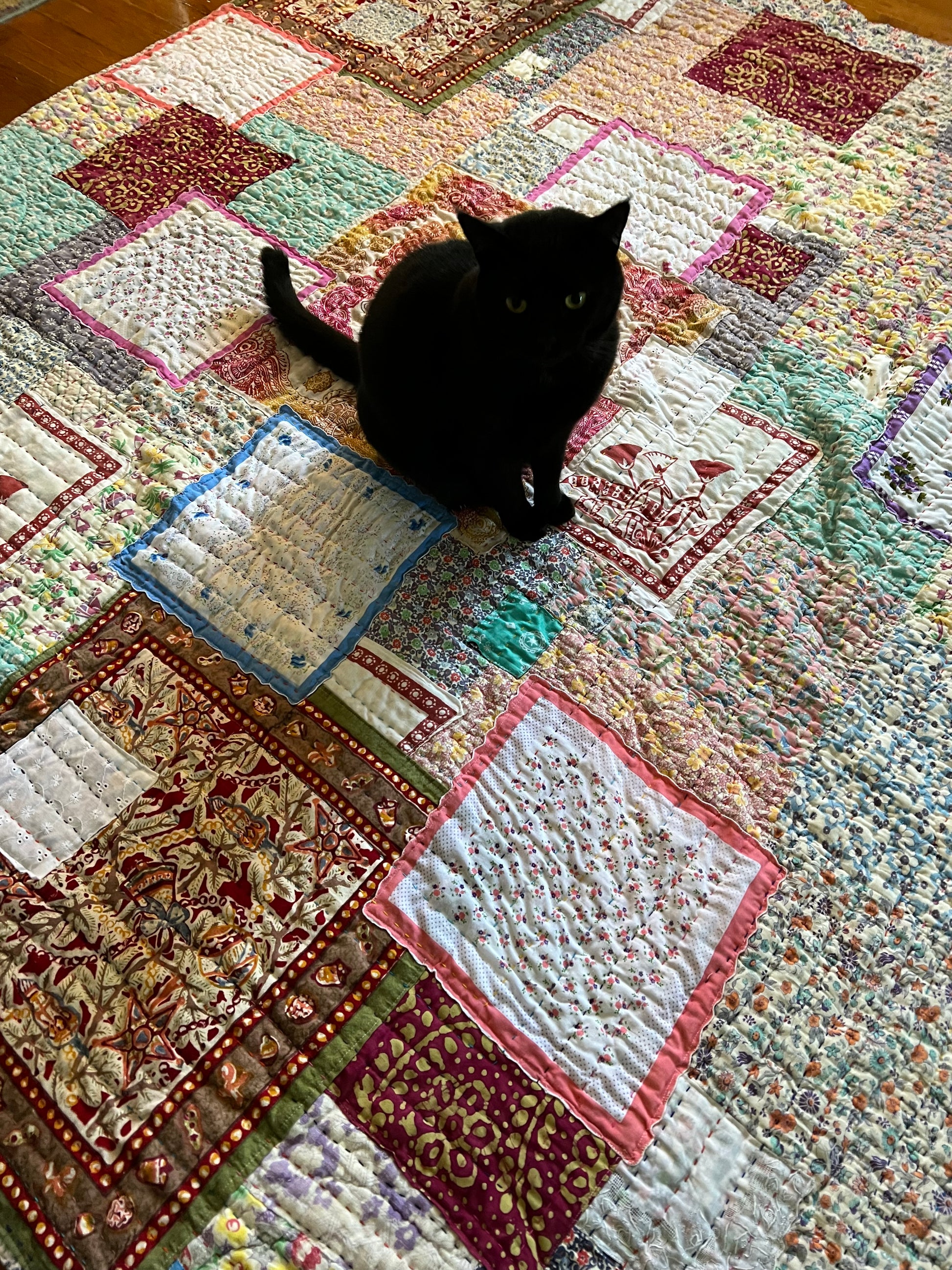 a black cat sits in the middle of the quilt, side/aerial view