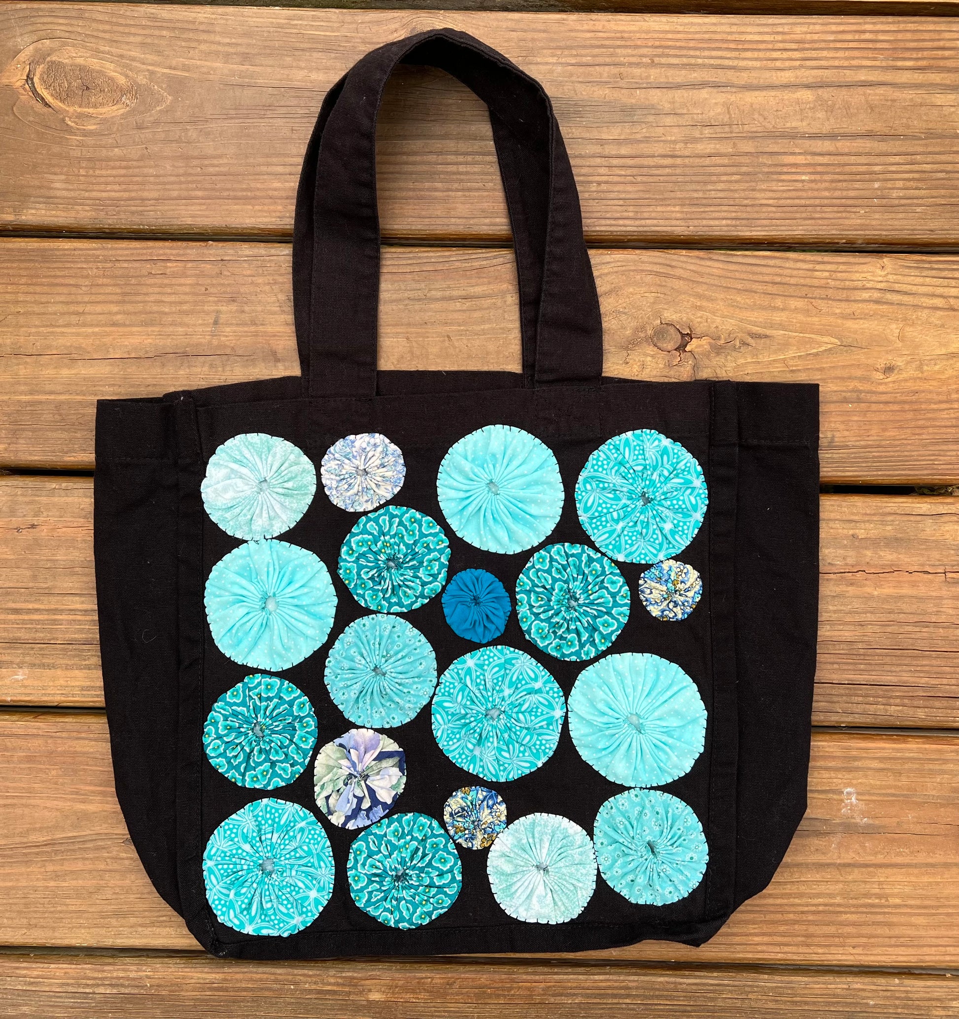 black tote bag with light blue yoyo circles sewn on top, aerial view against a wood deck background