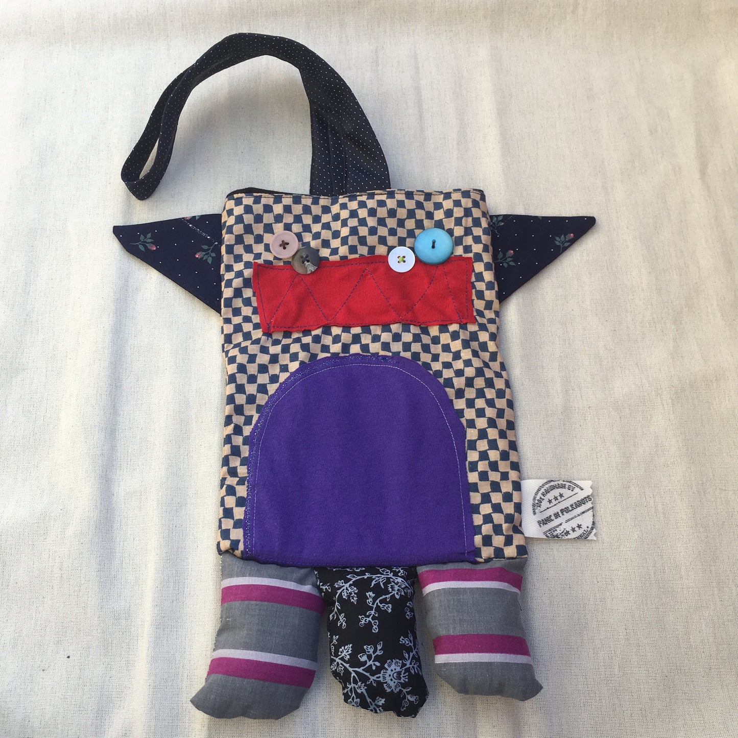 Monster tote bag, with necktie strap. Body is wavy checkered, and a purple tummy. a bright red mouth, and multiple button eyes. three legs on the bottom, and a set of antlers on either side of the head.