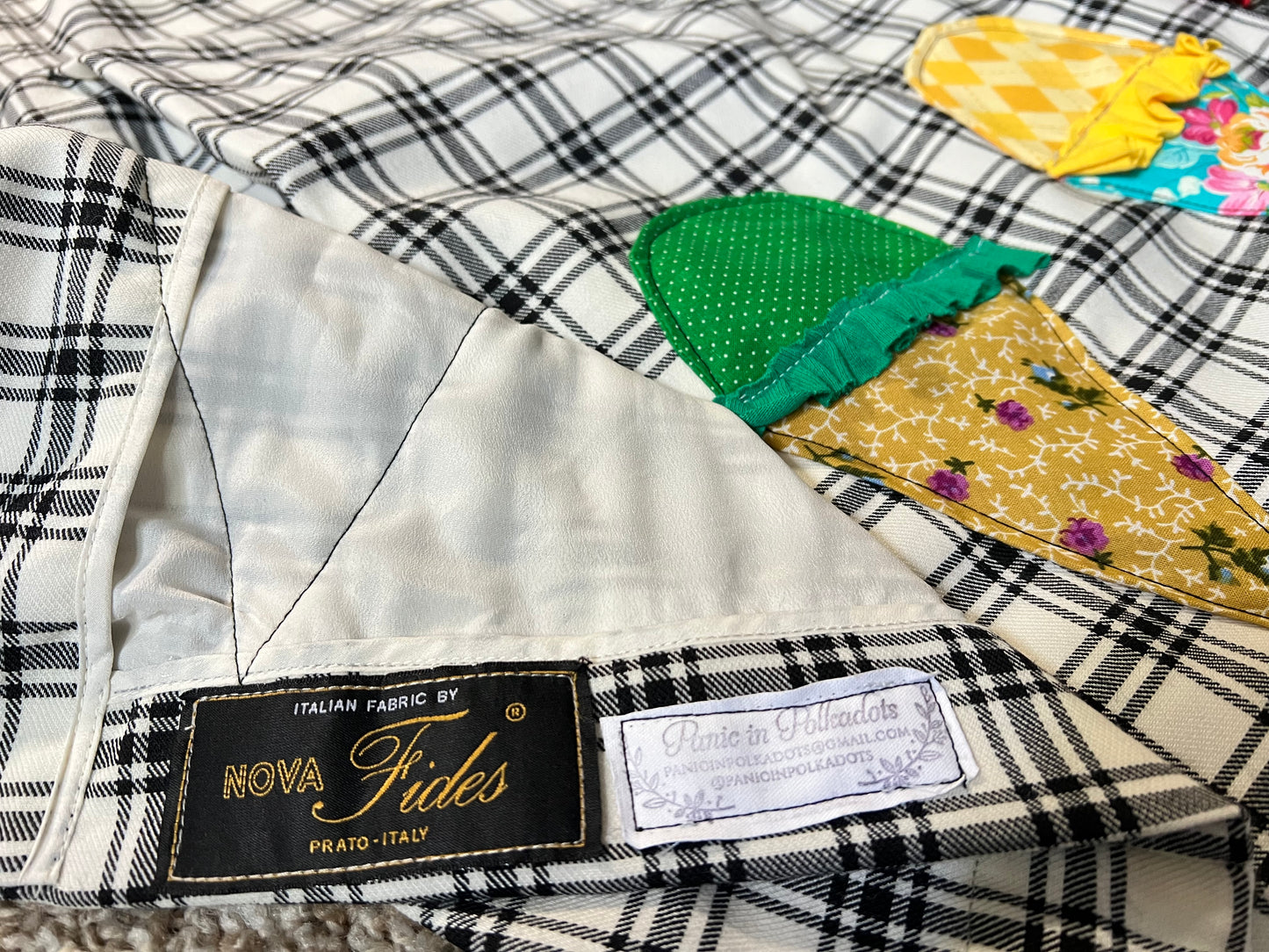 Close-up of a handmade fabric skirt featuring a black and white plaid pattern, a cream lining, and colorful applique details resembling ice cream cones. Two brand labels are visible: 'NOVA Fides' and 'panic in Polkadots'.