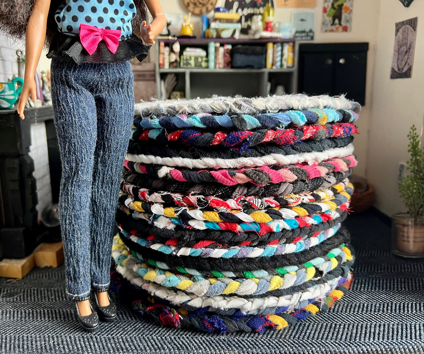 Stack of colorful braided rugs with a doll in a polka dot top and jeans standing next to them.
