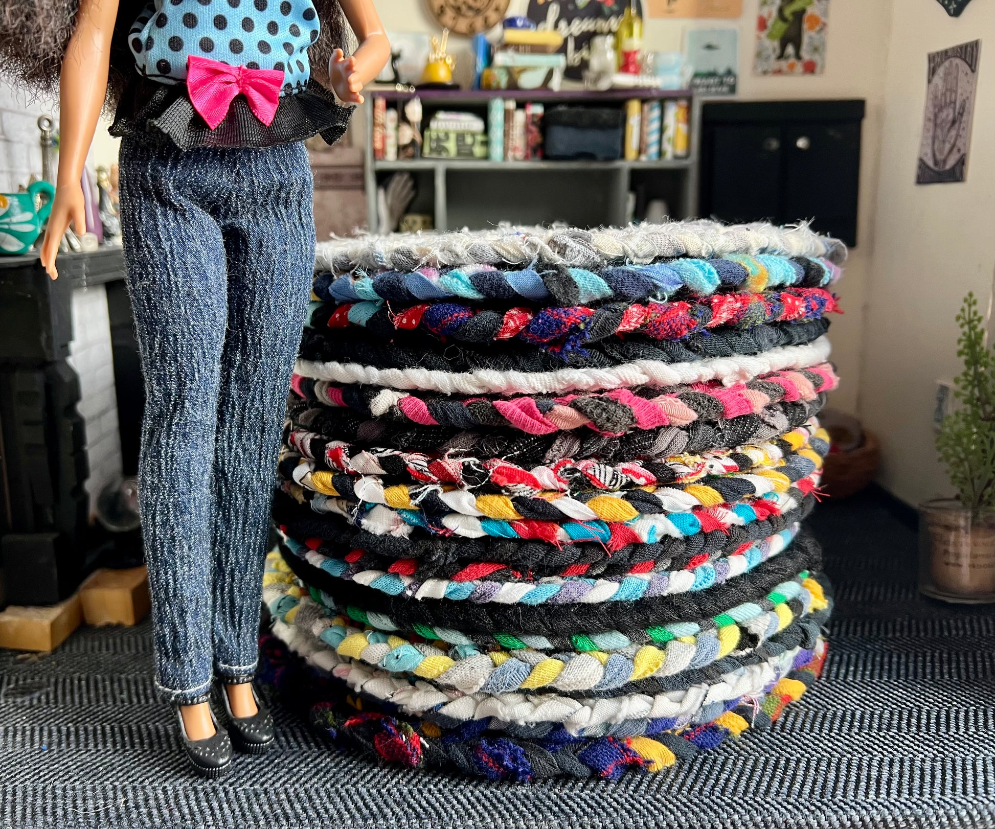 Stack of colorful braided rugs with a doll in a polka dot top and jeans standing next to them.