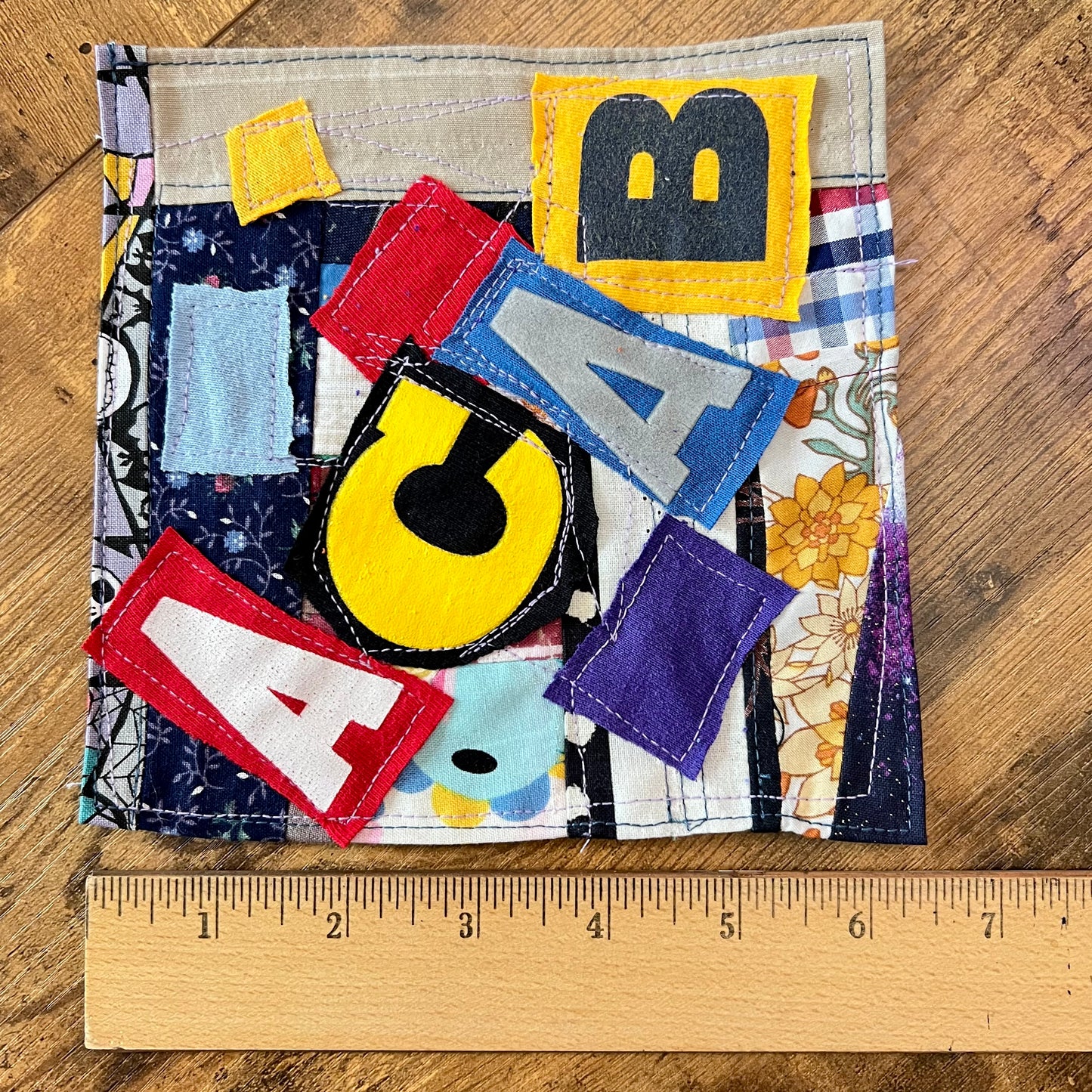 A handmade fabric patch laid flat on a wooden surface, with a wooden ruler along the bottom edge for scale. The patchwork design features various colorful letters and patterned fabric panels in yellow, blue, red, purple, black, and white.