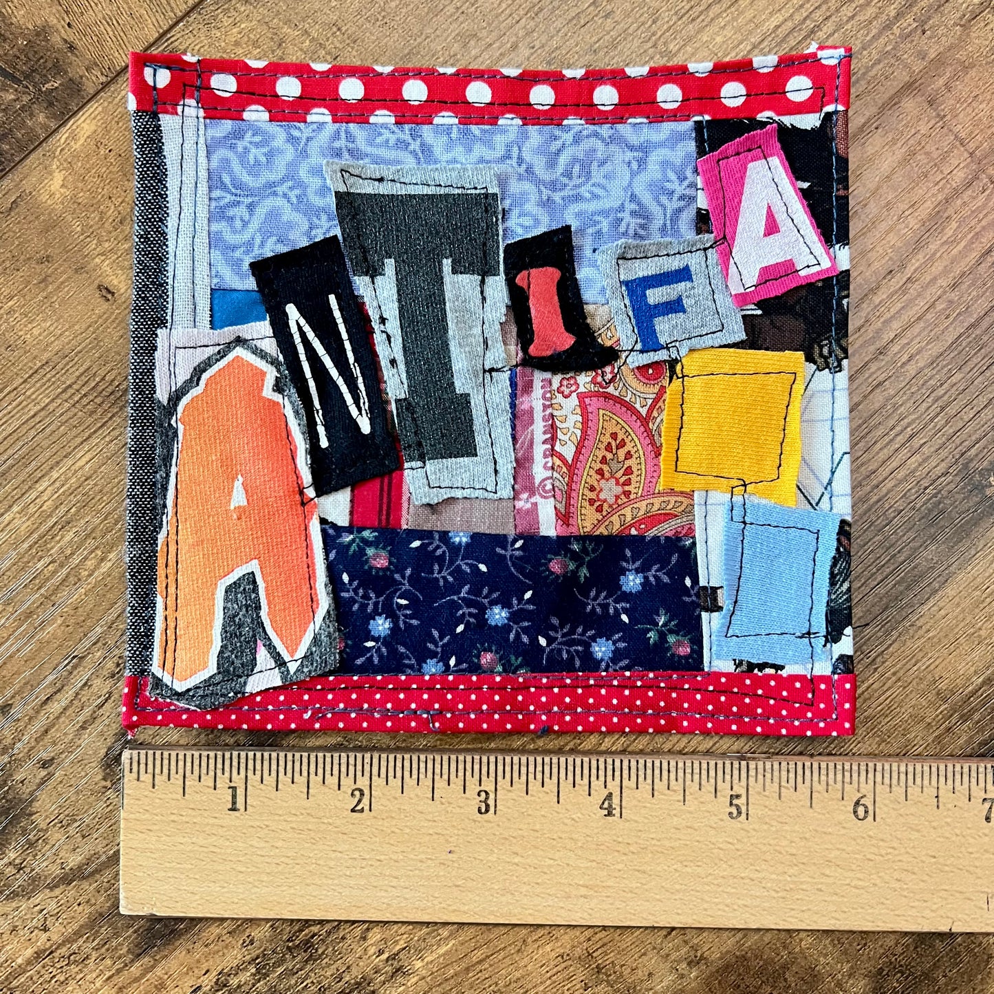 A handmade fabric patchwork letter quilt or pillow featuring letters A, N, I, F, and L stitched onto various patterned fabrics, with red and white polka dot trim, shown on a wooden surface with a wooden ruler for scale.