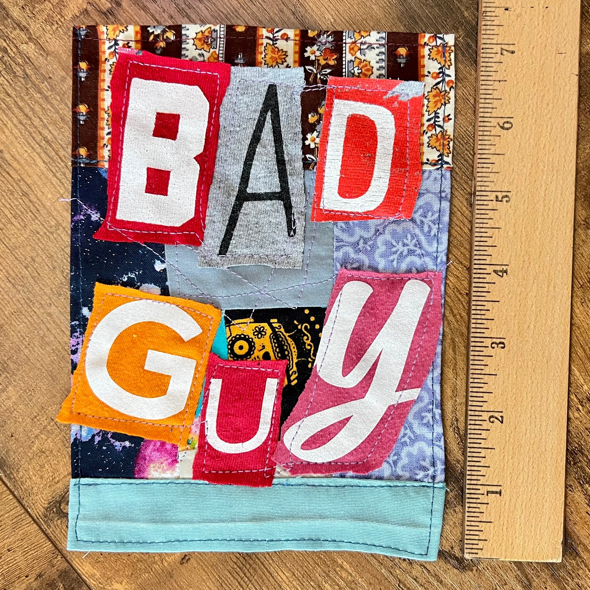 A handmade fabric patch spelling out 'BAD GUY' in bold letters, displayed on a wooden surface next to a wooden ruler for scale.
