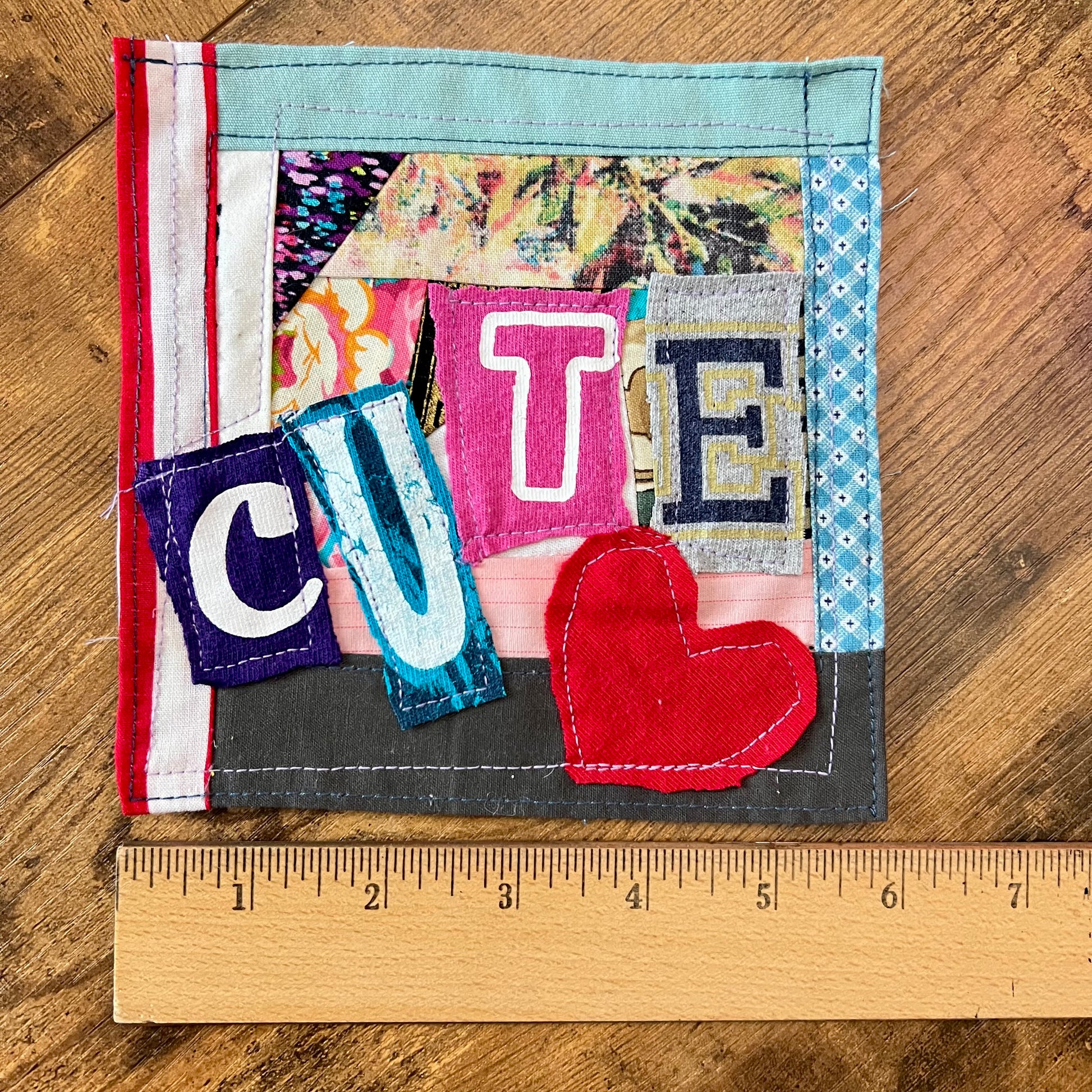 A handmade fabric square with patchwork lettering spelling 'CUTE' and a red heart applique, shown on a wooden surface with a wooden ruler below for scale.