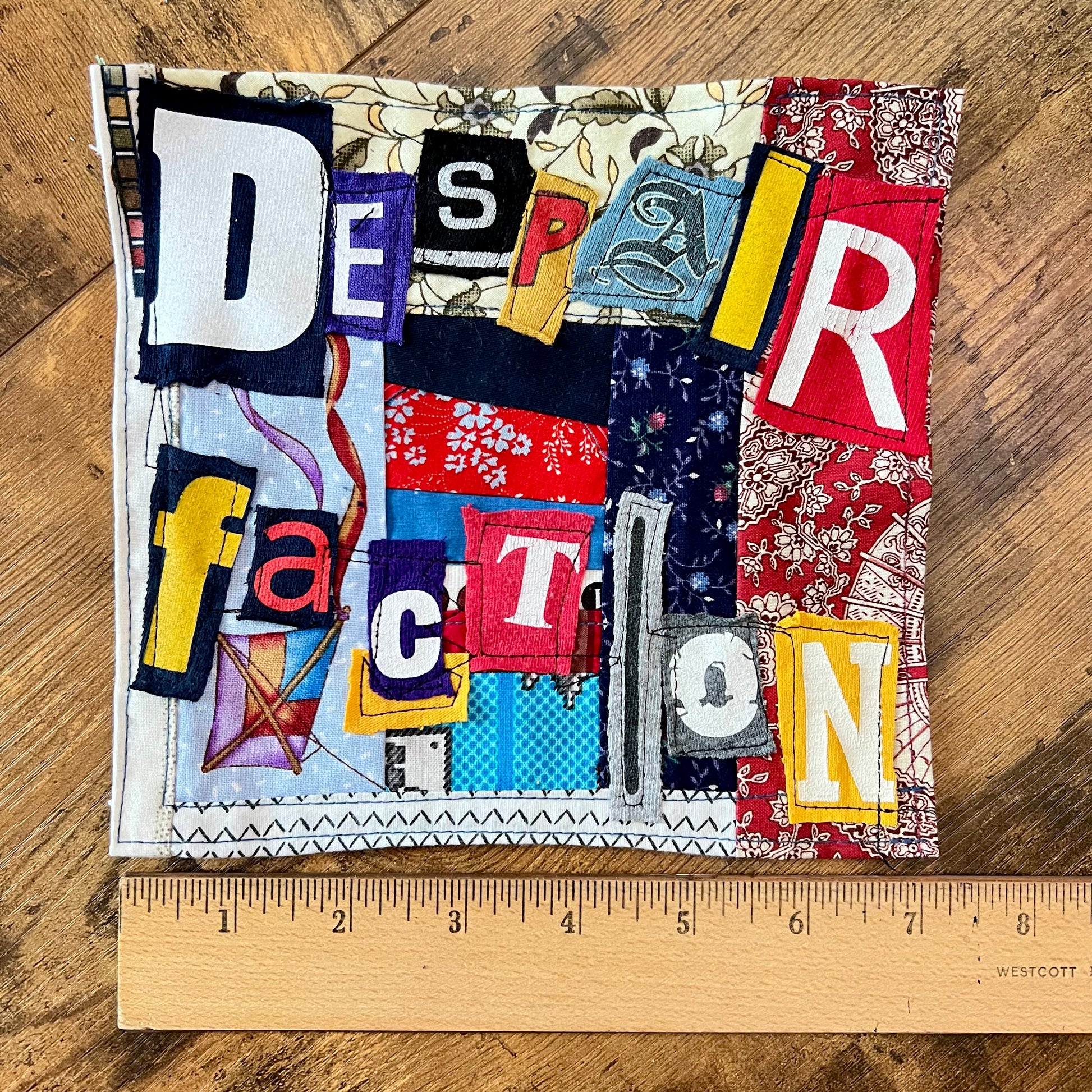 A square handmade fabric patch spelling out 'DESPAIR' and 'ACTION' using colorful cut letters on a patterned background, displayed on a wooden surface with a wooden ruler underneath for scale.