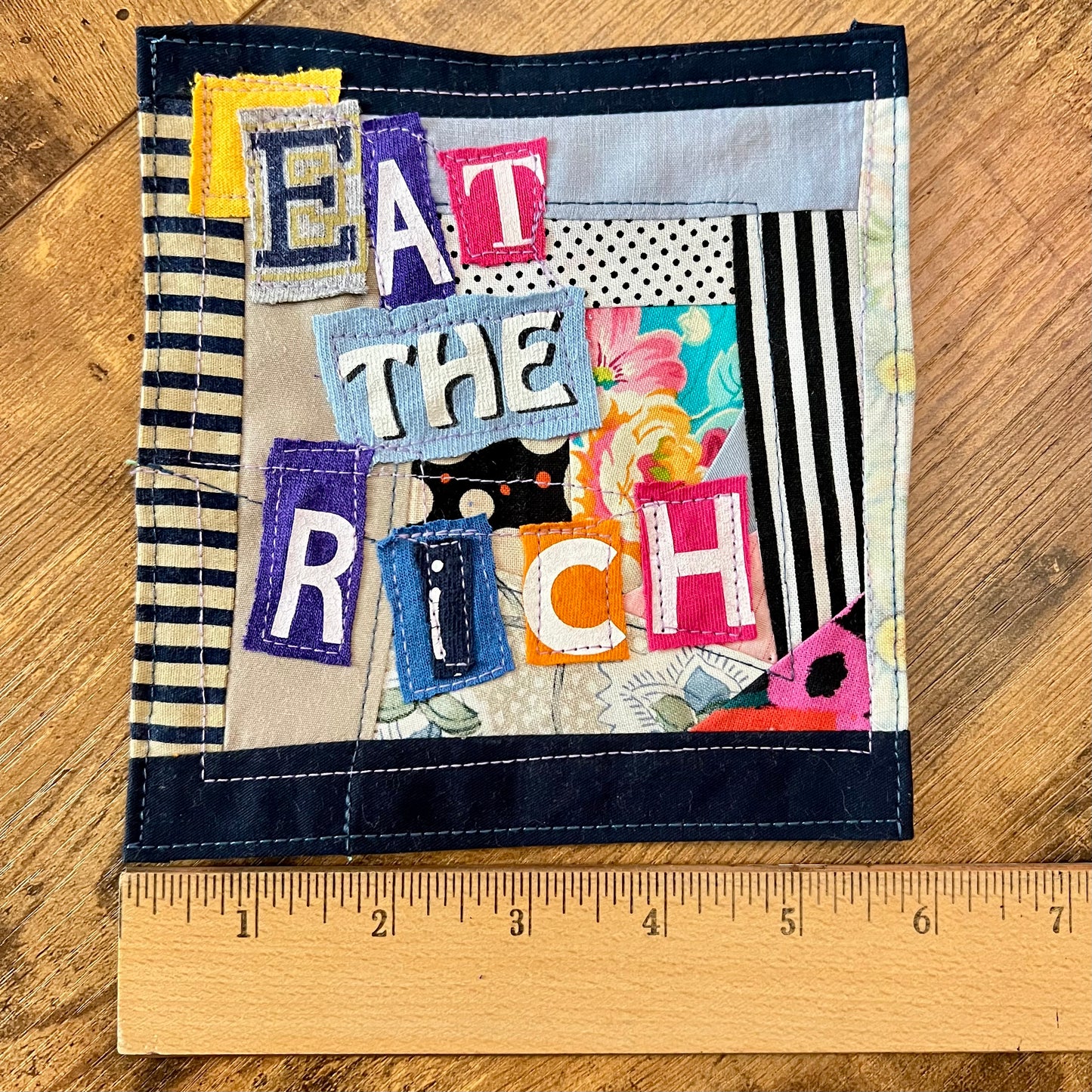 A square handmade fabric patch with the phrase 'EAT THE RICH' made from colorful fabric letters and patterned panels, shown on a wooden surface with a ruler at the bottom for scale.