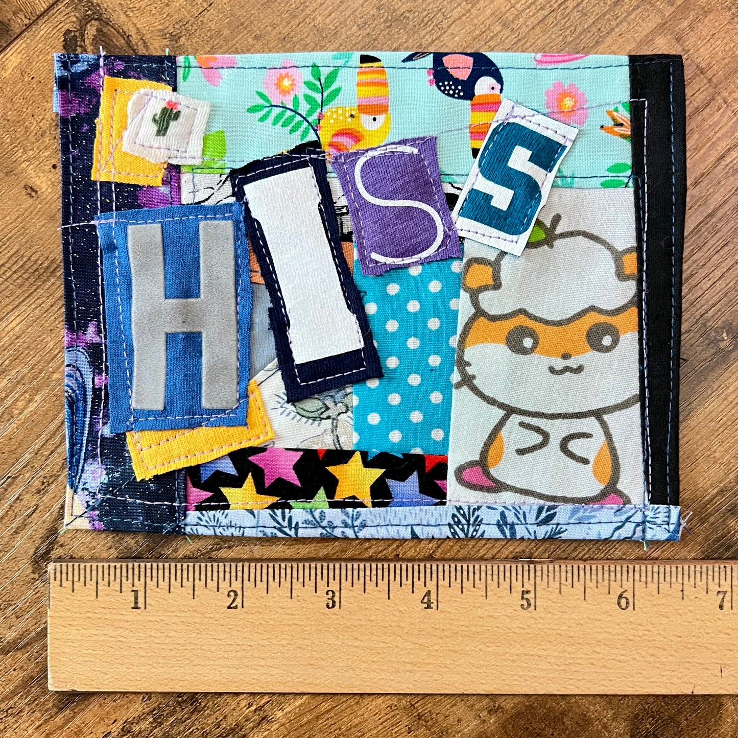 A handmade fabric patch spelling 'HIS' in patchwork fabrics, shown on a wooden surface with a wooden ruler underneath for scale. The wallet features various colorful patterned patches, including blue polka dots, yellow stars, and a cartoon hamster.