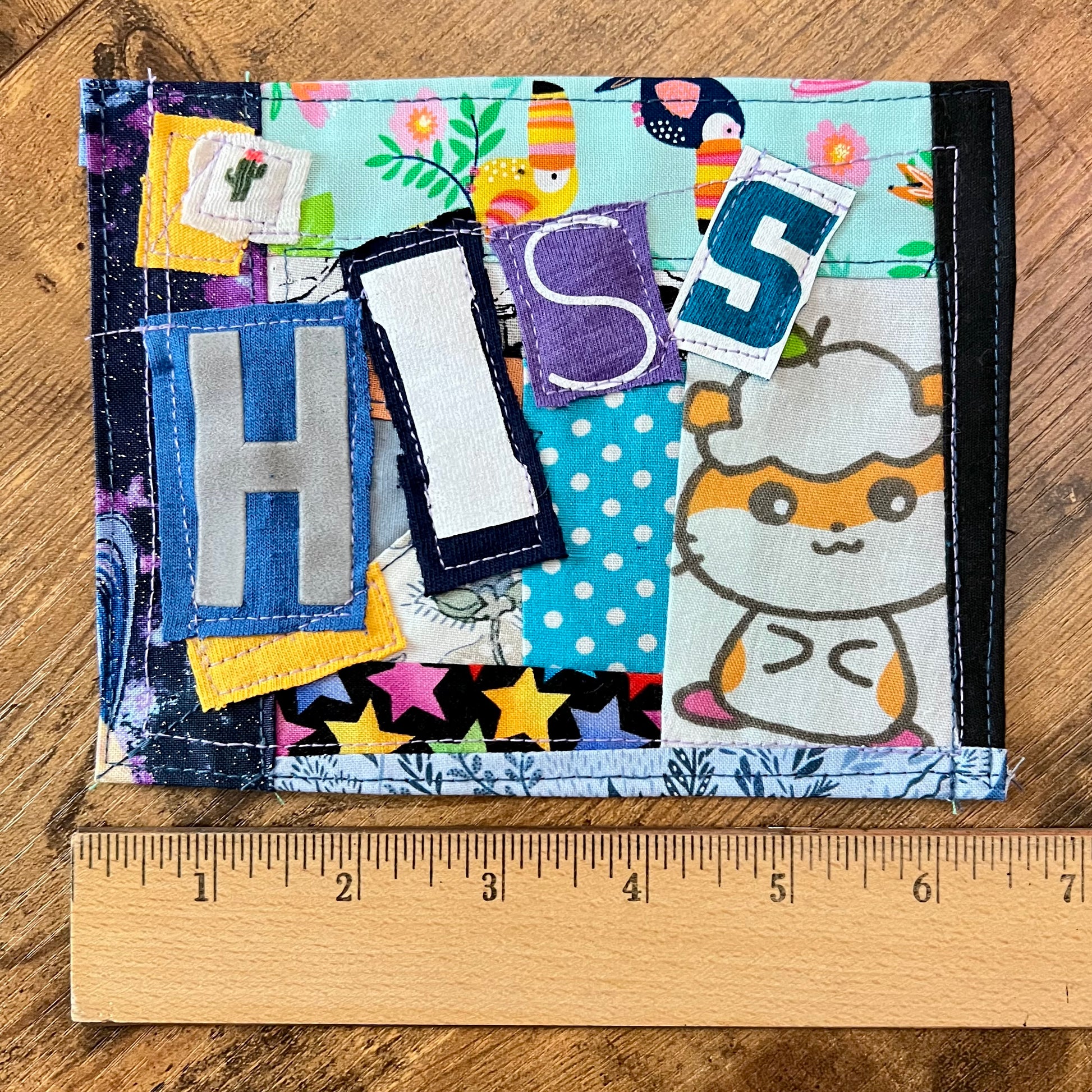 A handmade fabric patch spelling 'HIS' in patchwork fabrics, shown on a wooden surface with a wooden ruler underneath for scale. The wallet features various colorful patterned patches, including blue polka dots, yellow stars, and a cartoon hamster.