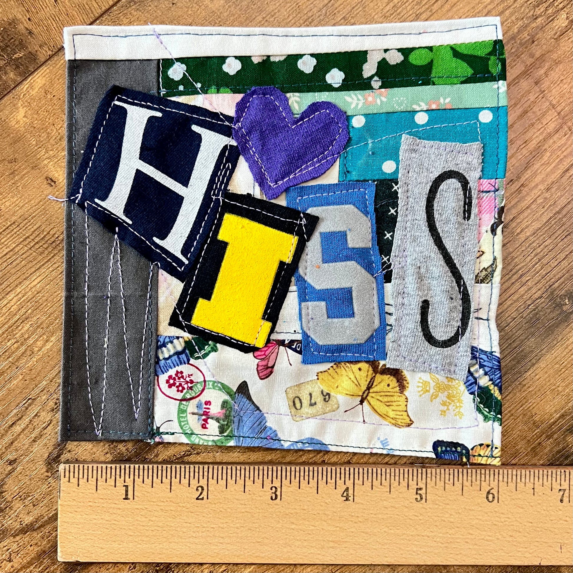 A handmade fabric patch or quilted lettering piece with letters H, I, S, and S, a purple heart, and butterfly details on a wood background with a wooden ruler at the bottom showing measurements.