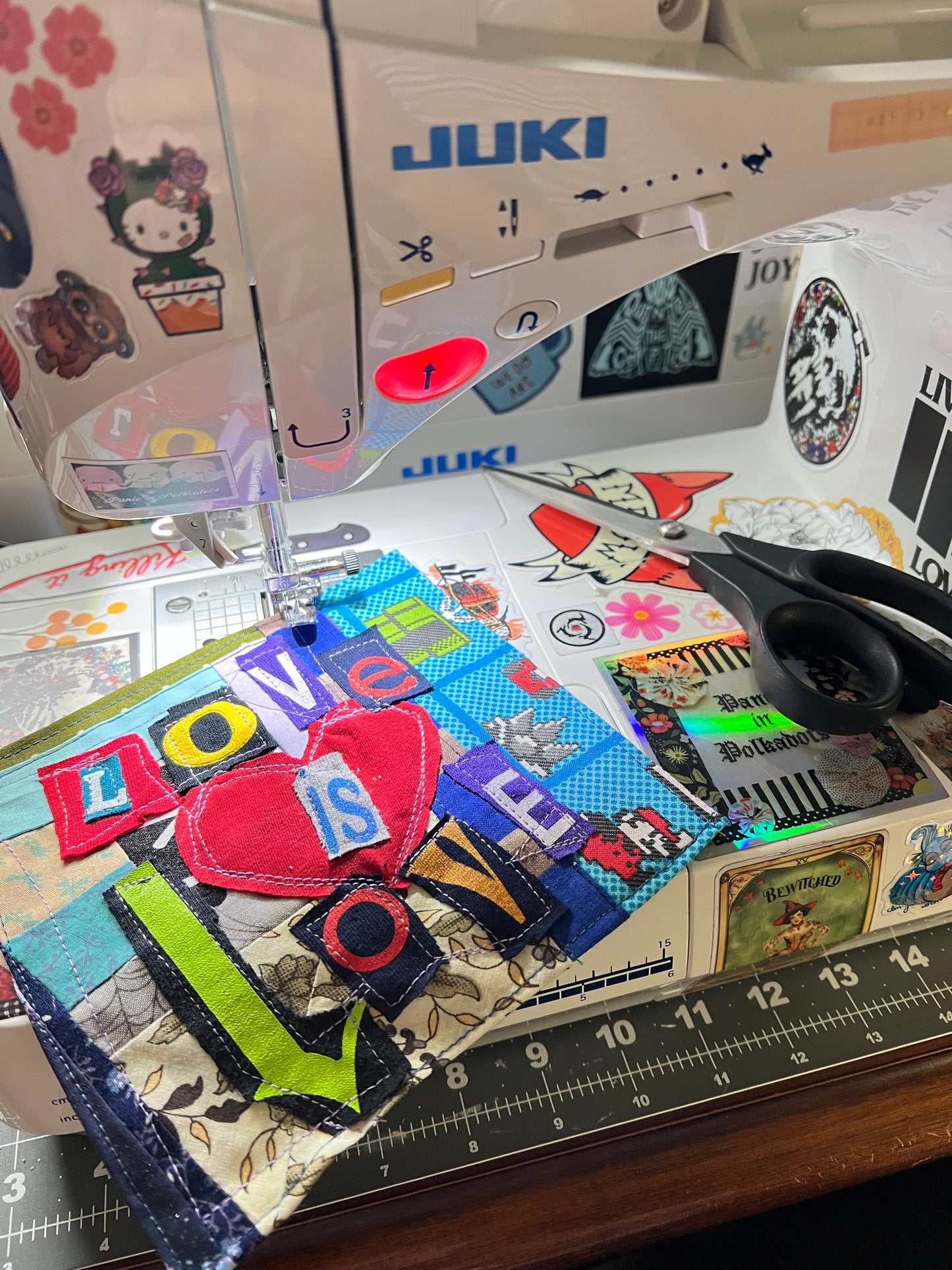 A close-up of a sewing machine with a colorful fabric patch under the needle, surrounded by sewing tools and a measuring ruler on a work surface.