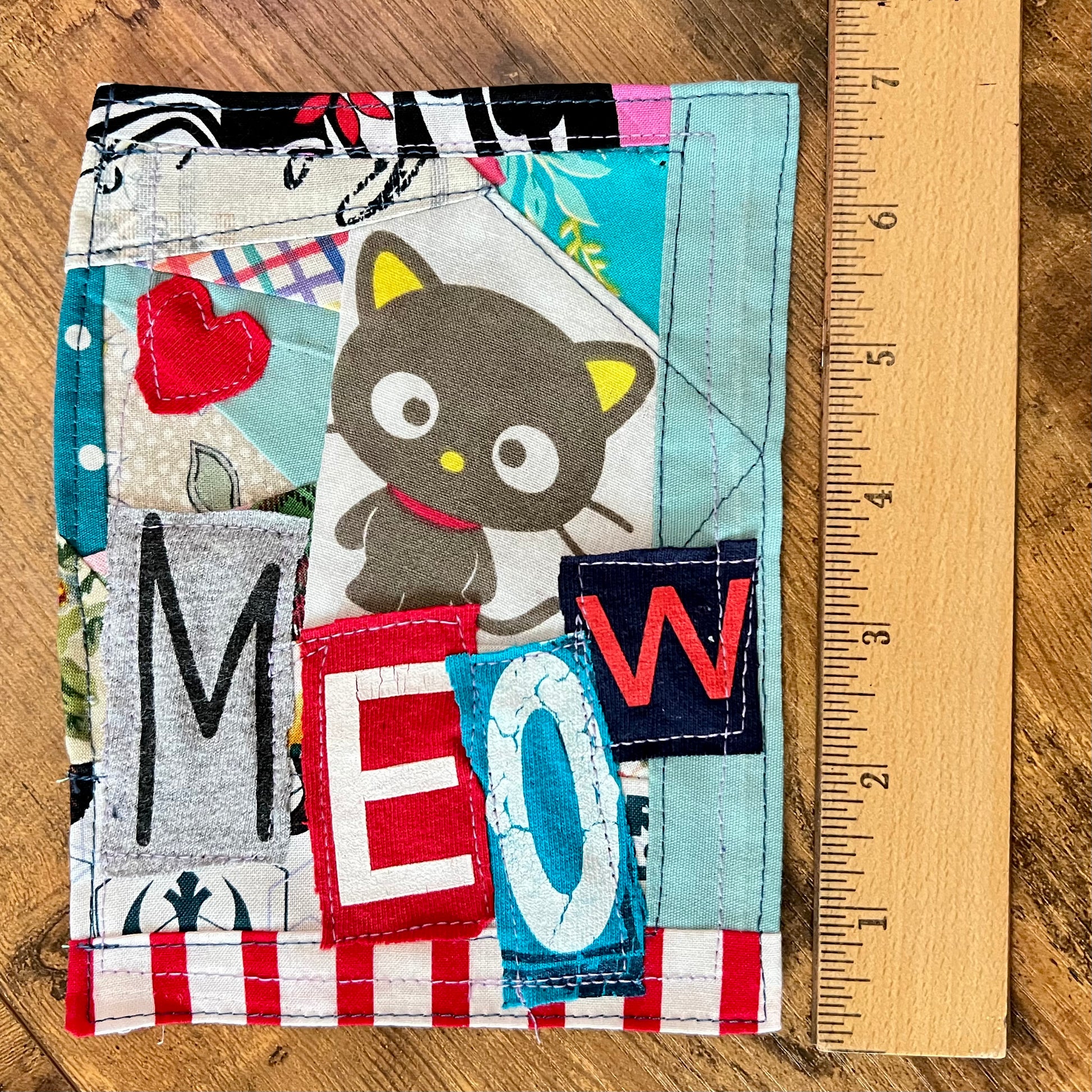 A handmade patchwork fabric patch featuring a cartoon cat, the word 'MEOW' in patchwork letters, and a red heart, shown next to a wooden ruler on a wooden surface.