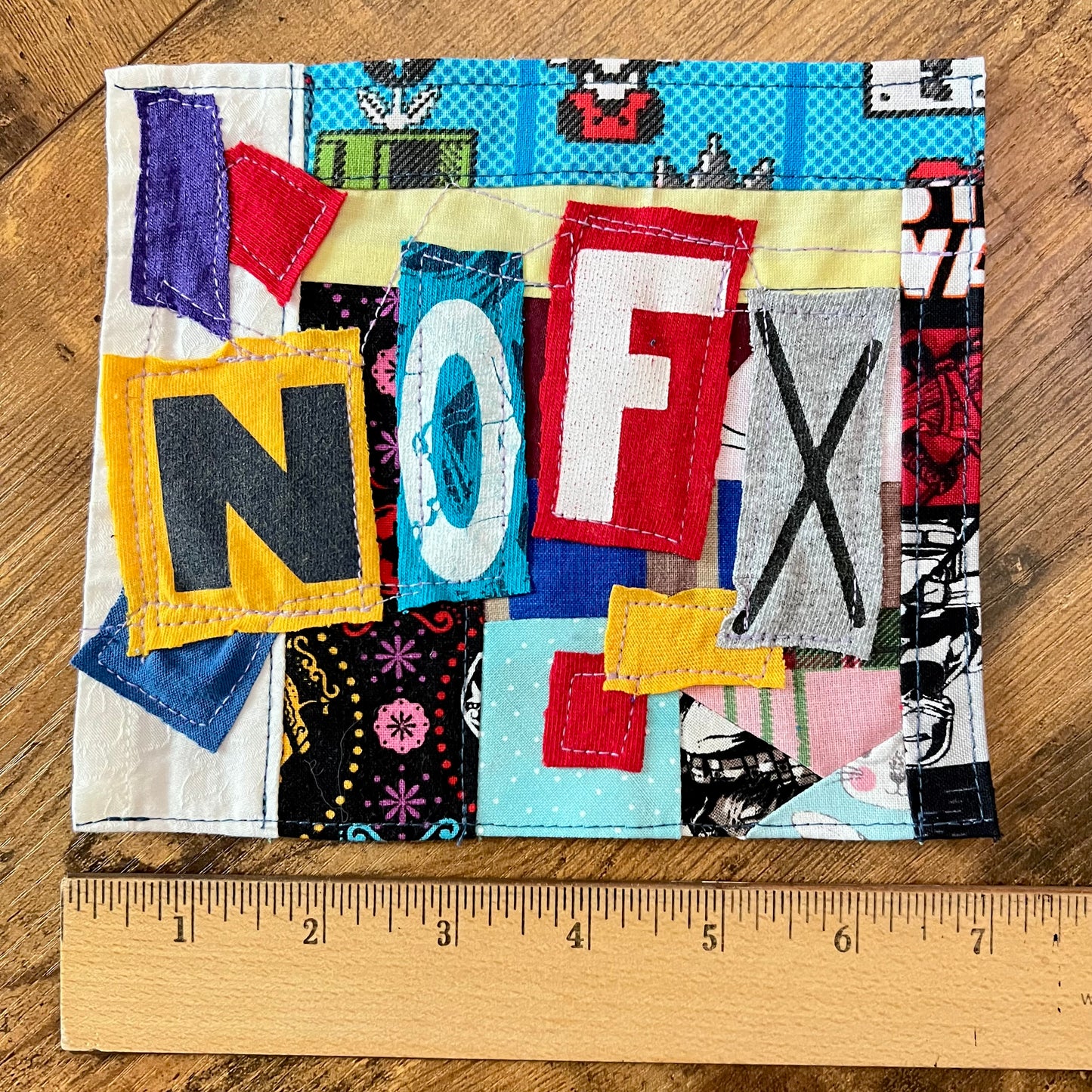 A handmade fabric patch spelling 'NOFX' on a wooden surface, with a wooden ruler at the bottom showing measurements.
