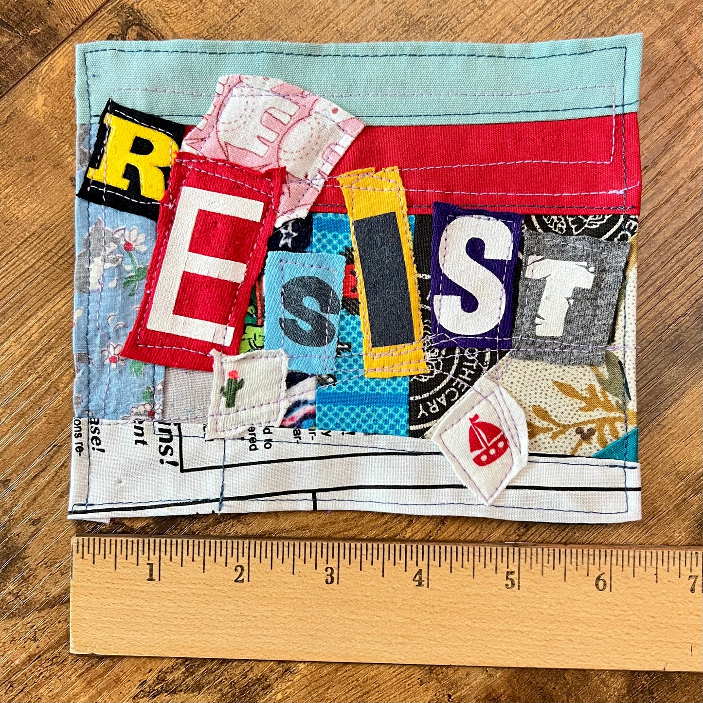 A square handmade fabric patch featuring the word 'RESIST' spelled out in colorful fabric letters, placed on a wooden surface with a wooden ruler at the bottom showing its size.