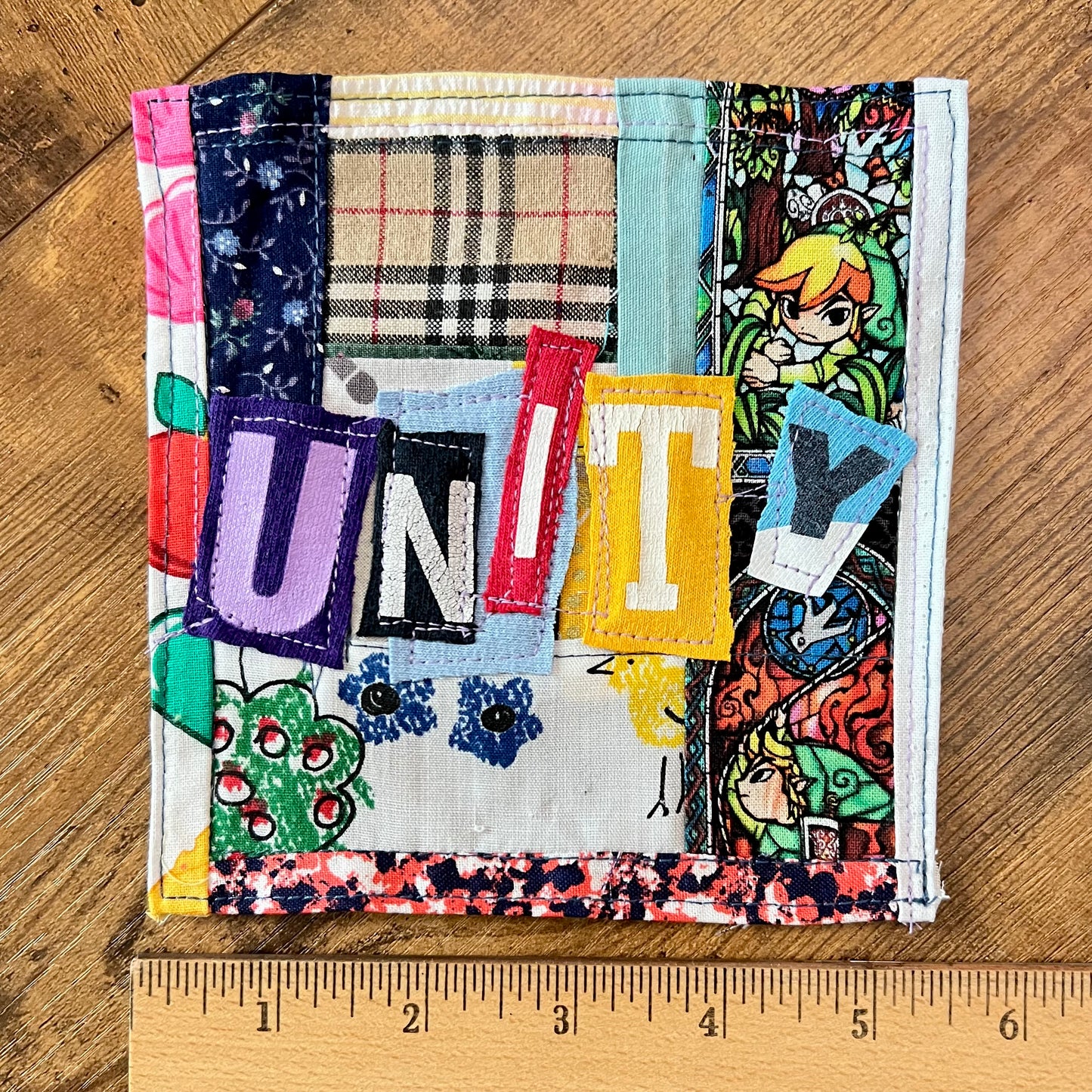 A square handmade patchwork fabric patch, featuring colorful stitched fabrics and tshirt letters spelling 'UNITY' and various patterned fabrics, photographed on a wooden surface with a wooden ruler at the bottom for scale.