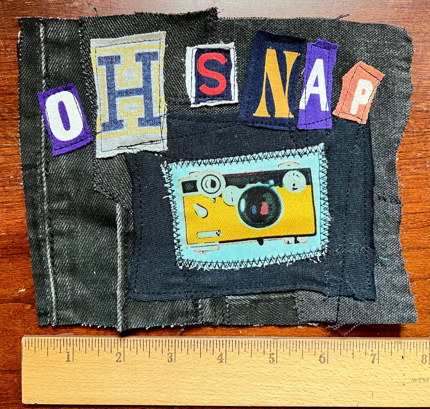 A handmade denim patch featuring the letters 'OH SNAP' in colorful patches and a yellow camera applique on a light blue background, displayed on a wooden surface with a ruler at the bottom for scale.
