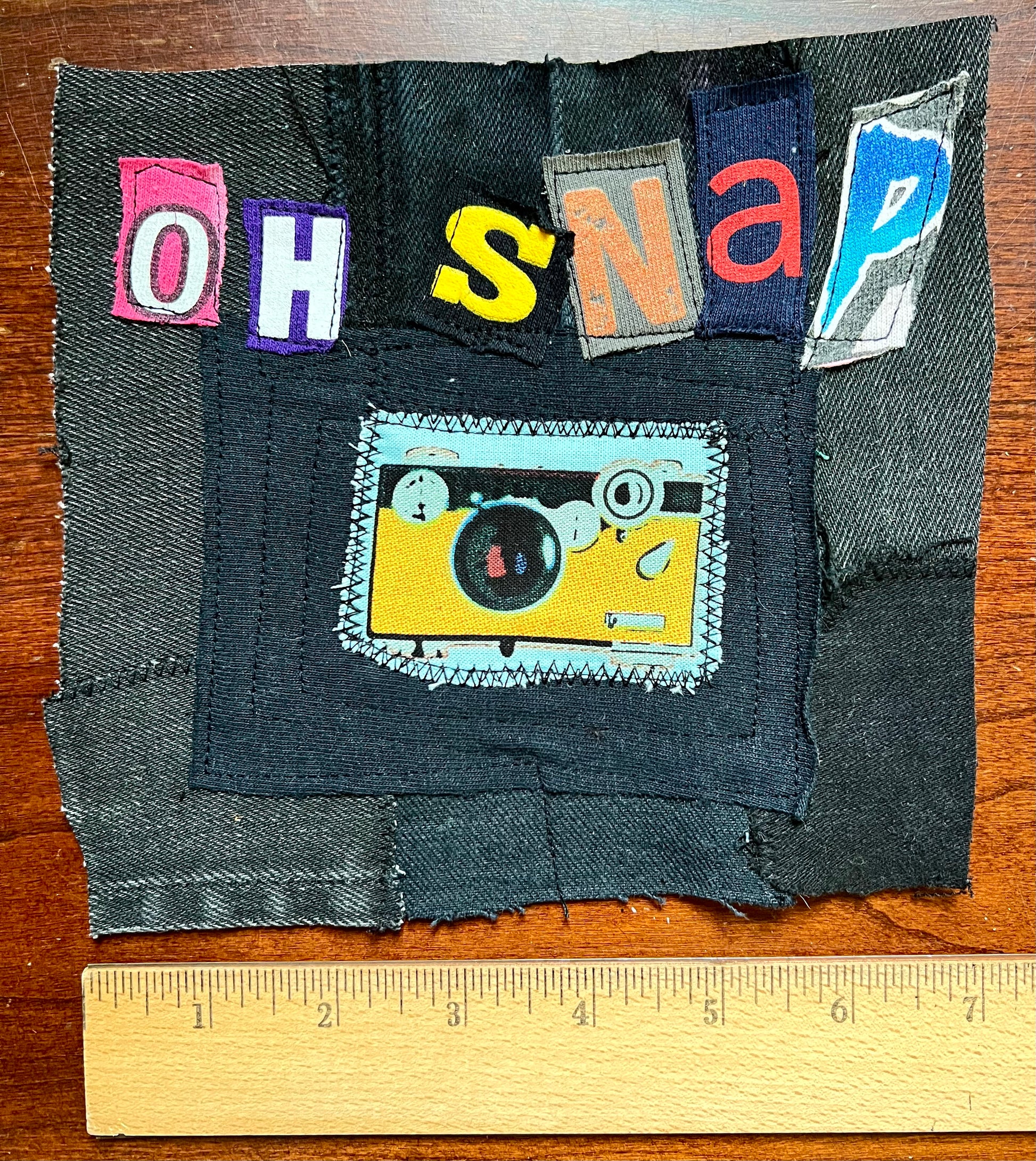 A handmade patch made from dark denim fabric with colorful letter patches spelling 'OH SNAP' and a yellow camera patch in the center, shown on a wooden surface with a ruler at the bottom for scale.