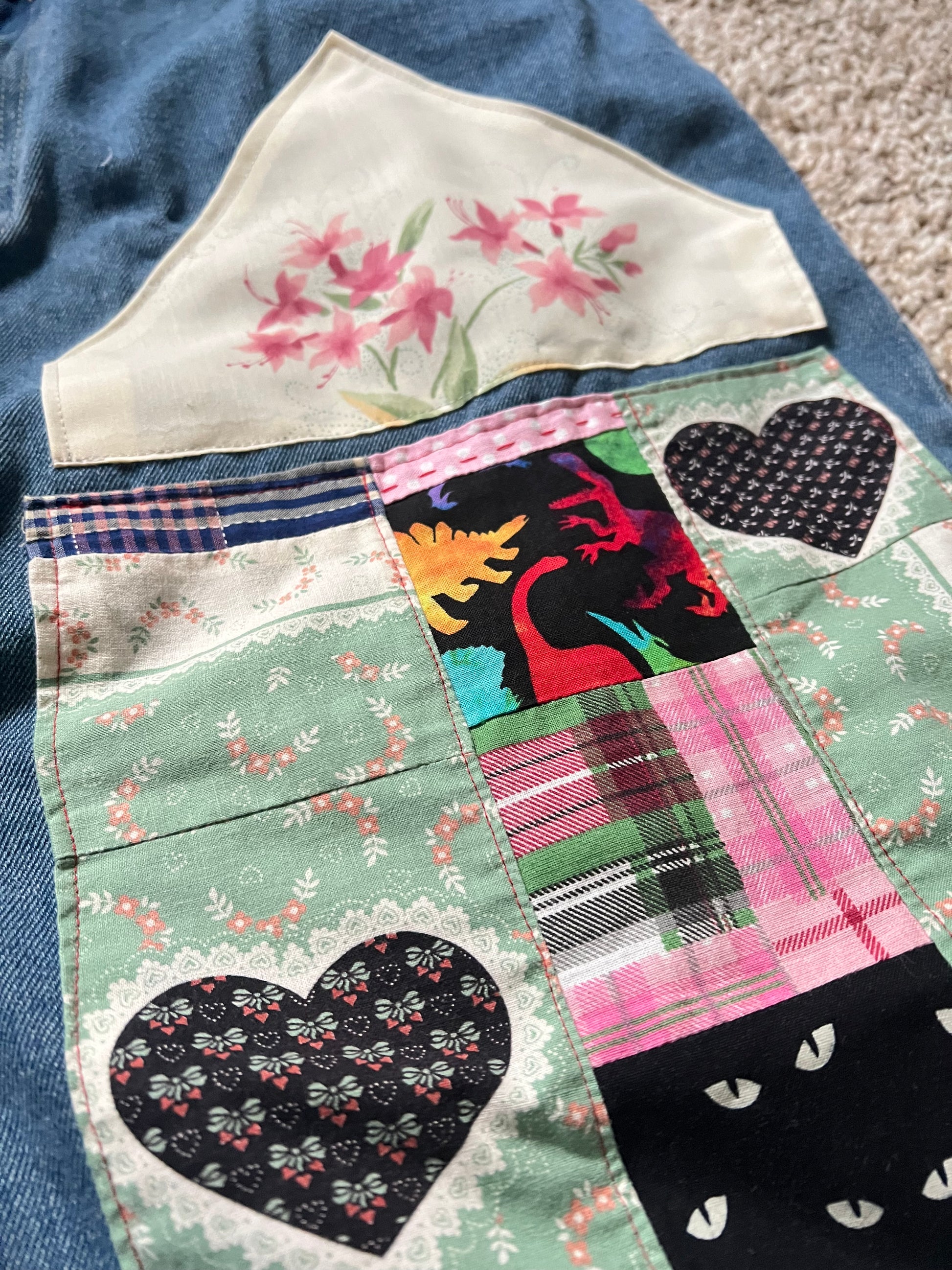 A close-up of a handmade patchwork quilt featuring various fabric patterns including floral prints, plaid, and heart-shaped motifs, laid flat on a blue denim fabric surface.