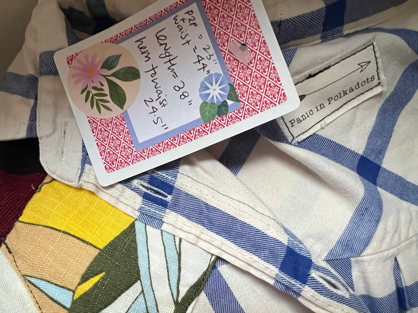 A close-up of a handmade patchwork blouse with a blue and white plaid pattern, featuring a colorful floral patchwork design on the front. A paper tag with handwritten measurements and a sewn-in label reading 'Panic in Polkadots' are attached.