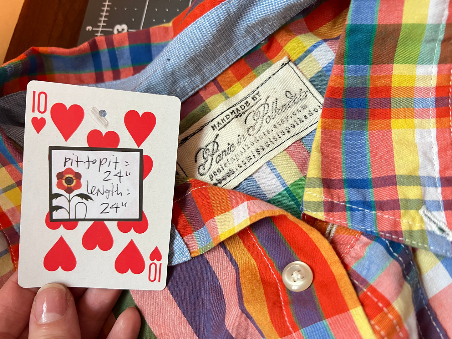 A close-up of a colorful plaid button-up shirt with a visible sewn label reading 'Handmade by Bento in Baltimore' and a hand holding a playing card over the front.