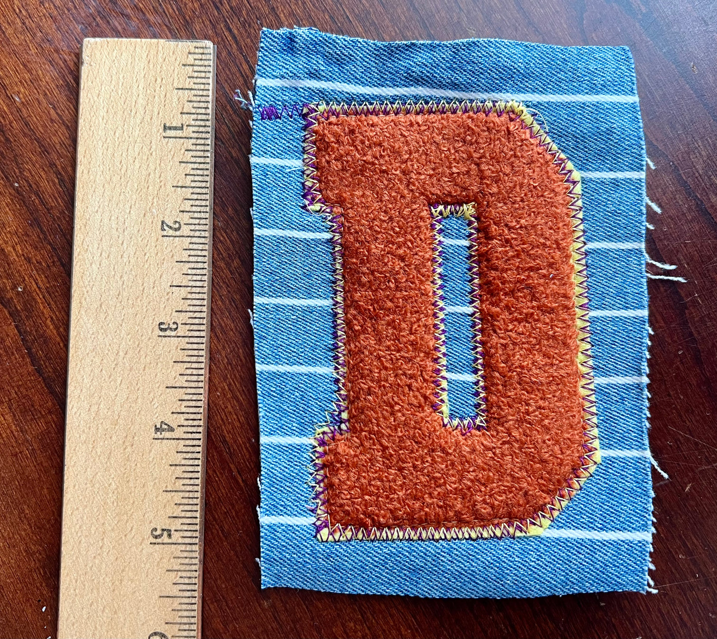Fabric patch with letter 'D' on a wooden surface next to a ruler