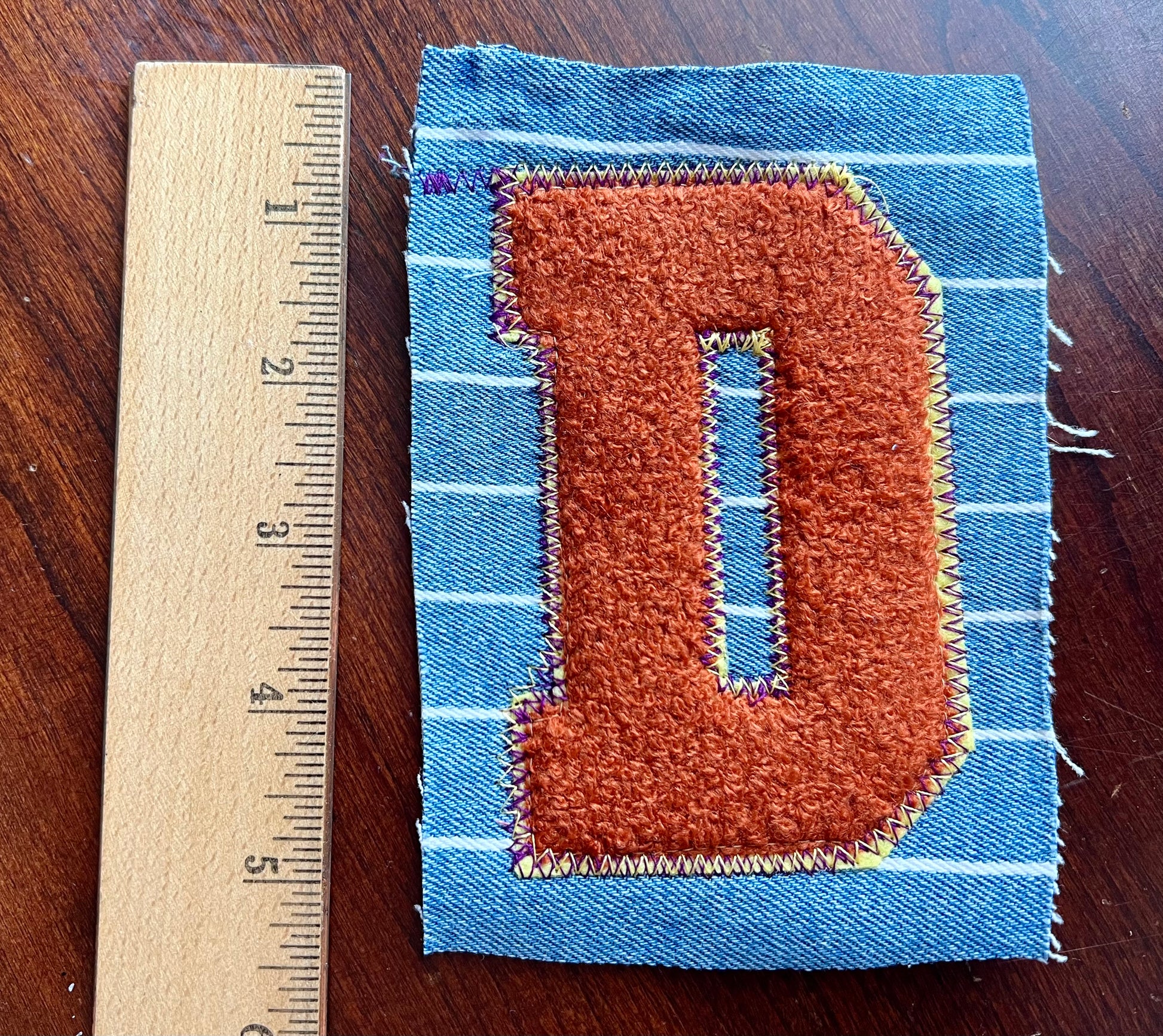 Fabric patch with letter 'D' on a wooden surface next to a ruler